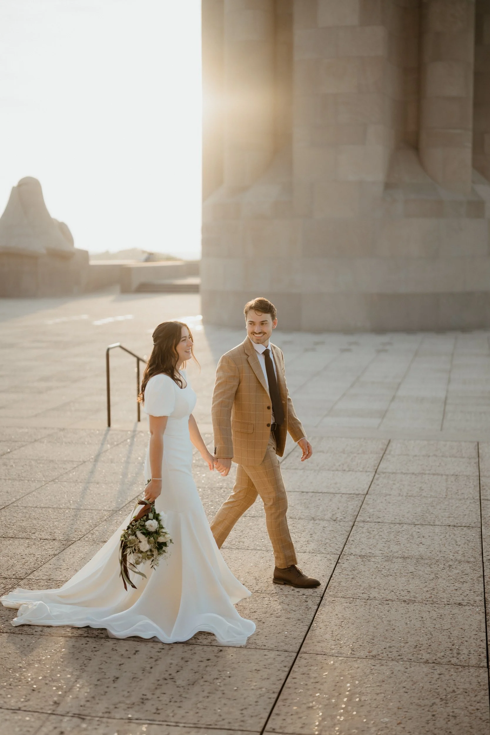 A newlywed couple walking hand-in-hand outdoors, the bride in a white wedding gown carrying a bouquet, and the groom in a tan checked suit, with a historic building in the background during sunset.