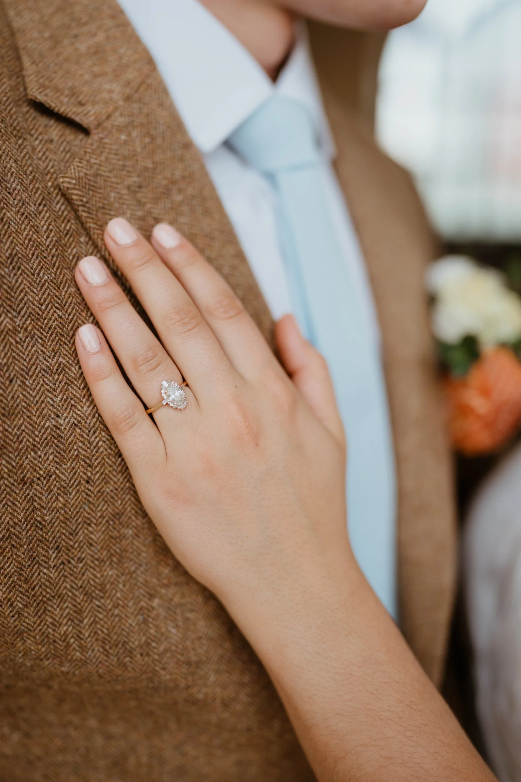 Close-up of a woman’s hand with a wedding ring resting on a man’s chest, woman wearing a brown jacket, man in a light blue shirt and tie.
