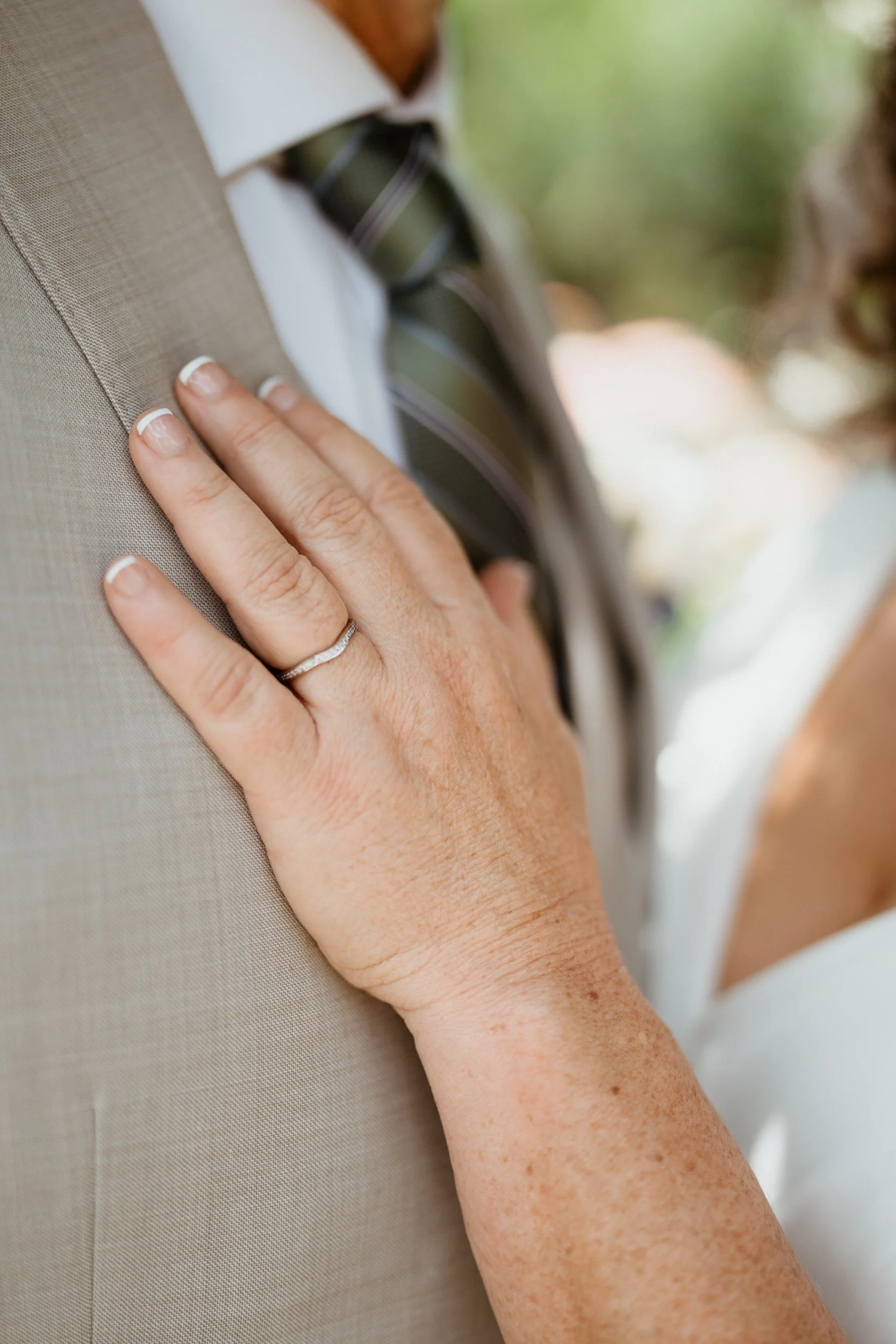 Close-up of a woman's hand with a wedding ring, resting on a man's beige suit jacket, during a wedding or special event.