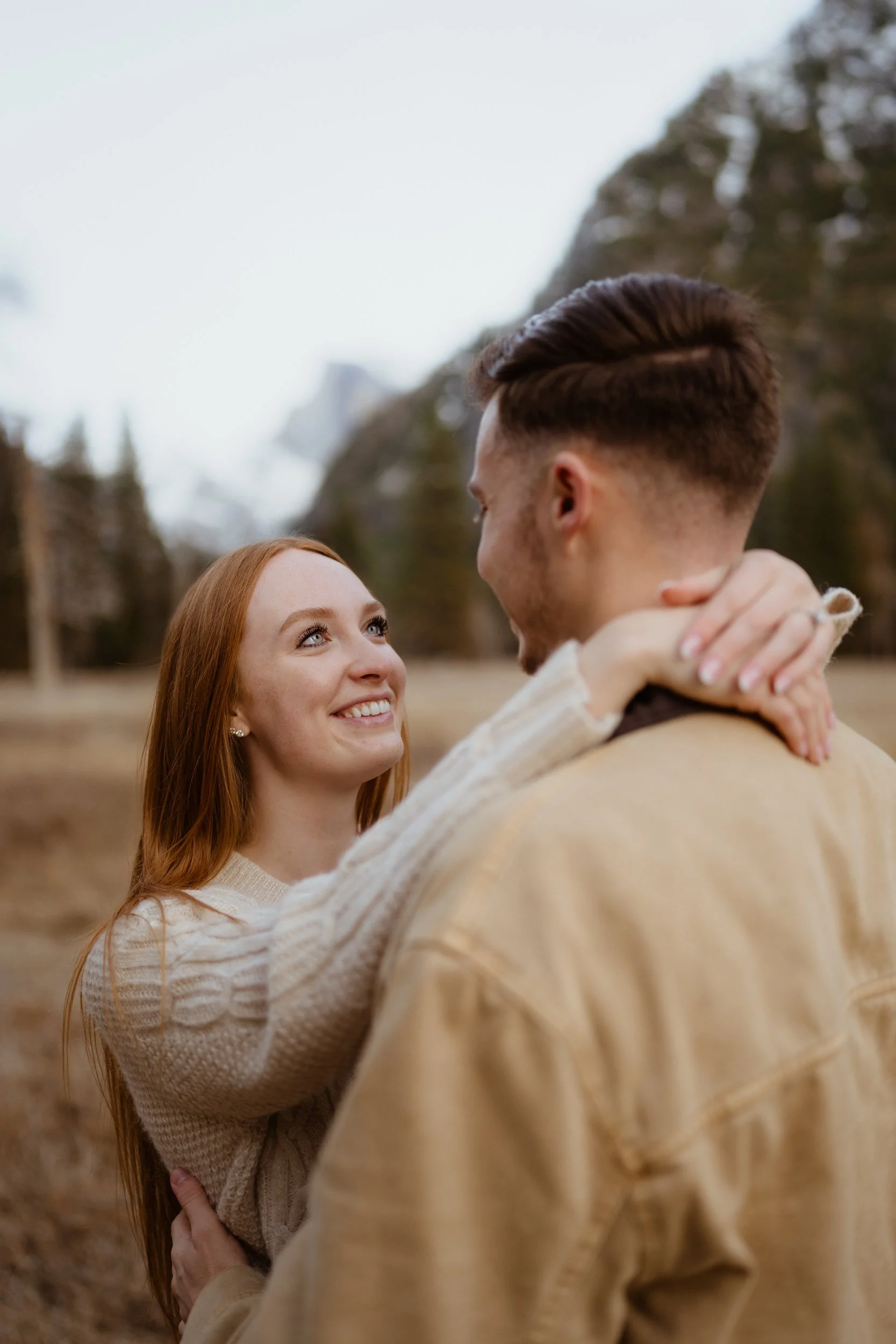 A woman and man embracing outdoors with mountains and trees in the background, smiling at each other.