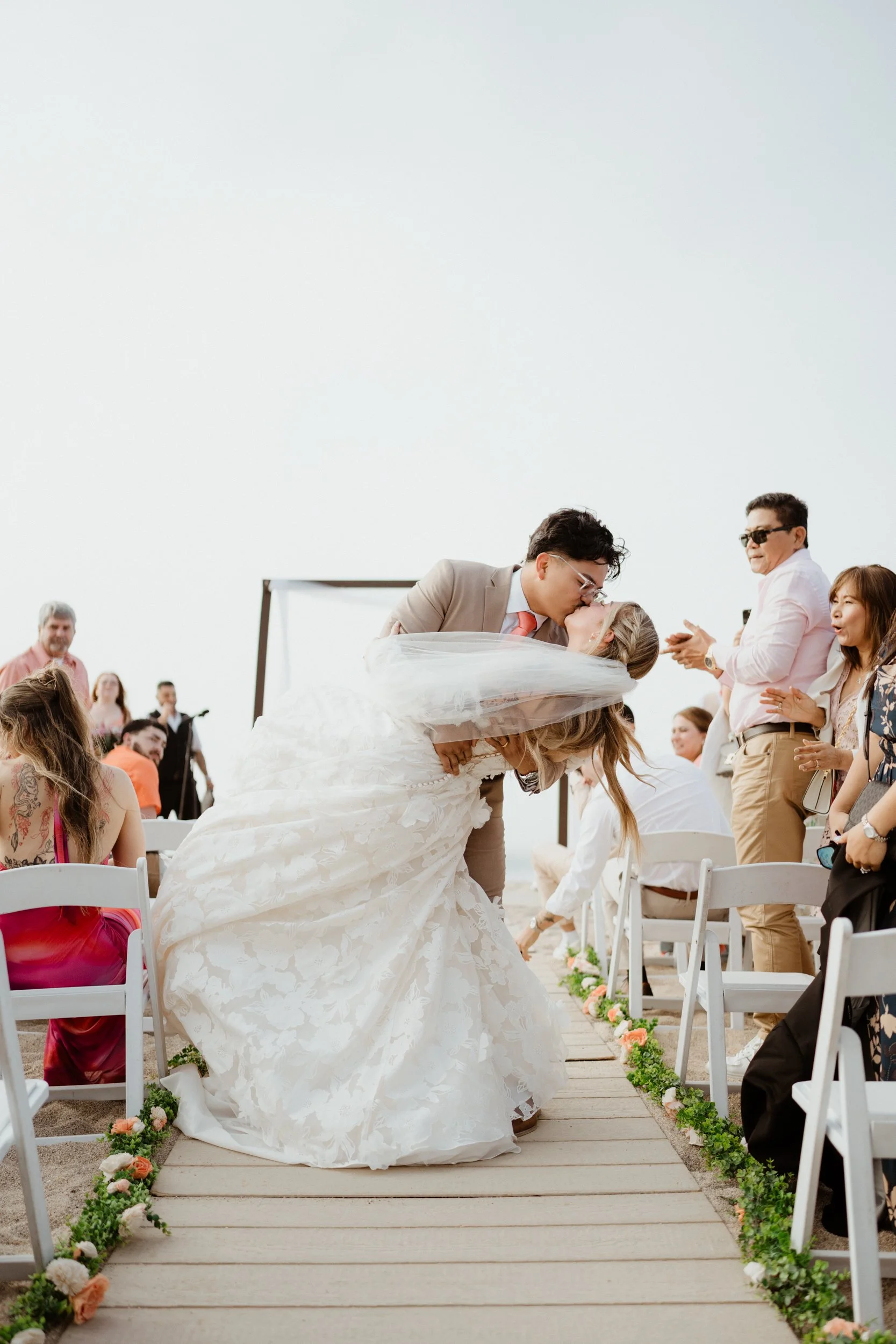 A newlywed couple sharing a kiss during their outdoor wedding ceremony on a wooden aisle decorated with flowers, with wedding guests clapping and smiling in the background.