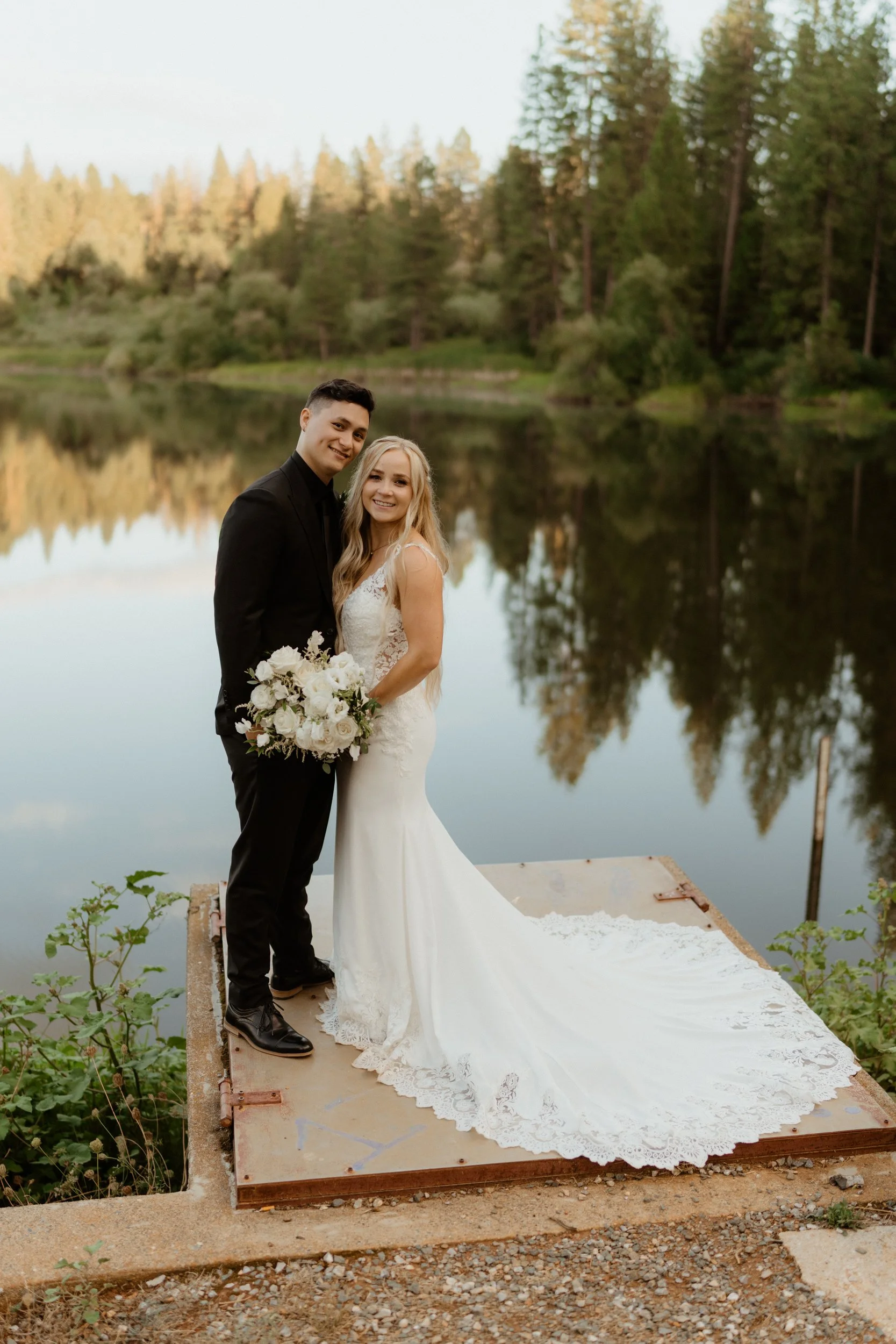 A newlywed couple in wedding attire standing on a dock by a calm lake surrounded by trees, smiling at the camera.