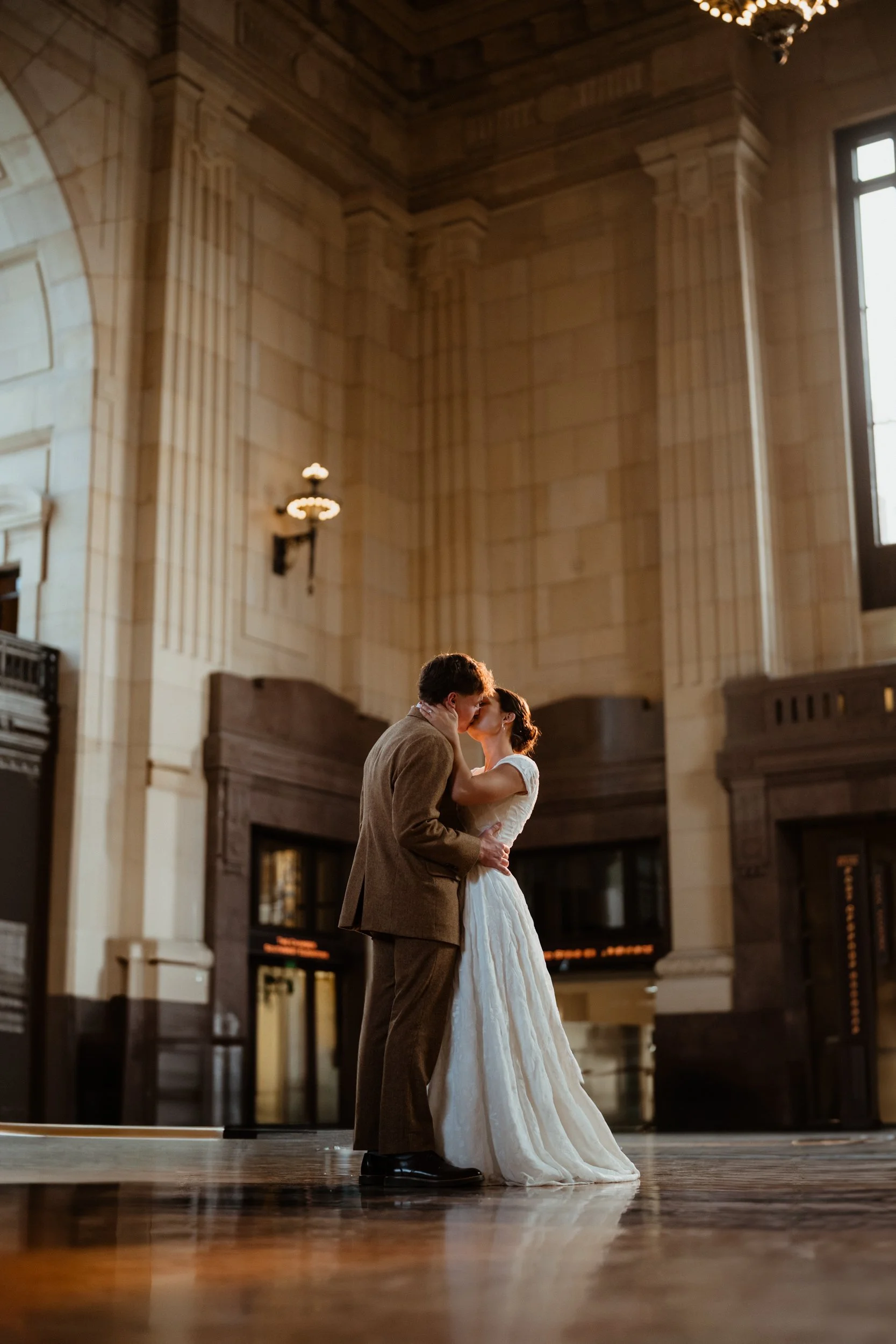A bride and groom sharing a kiss in a grand, historic train station with high ceilings, large windows, and vintage lighting.
