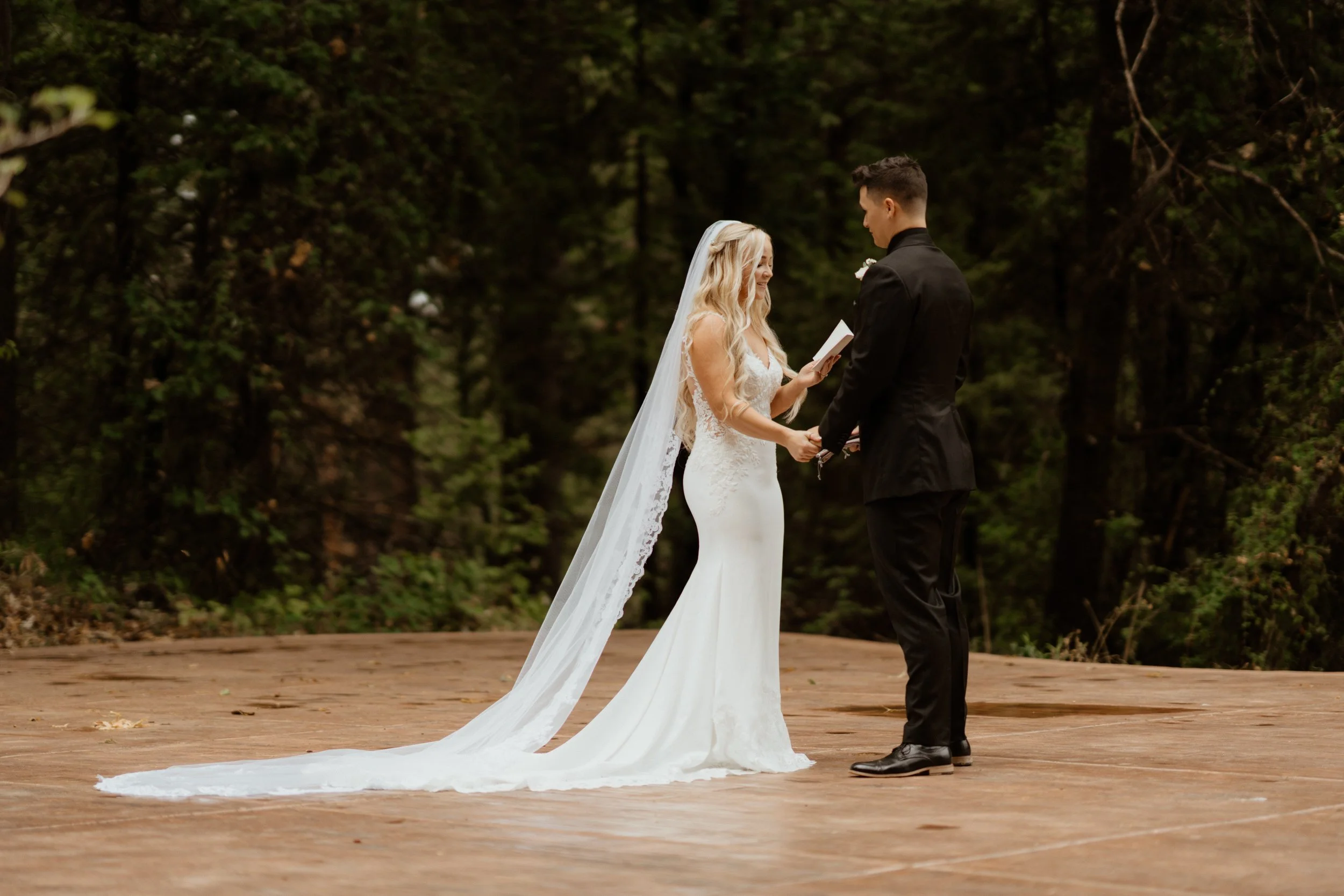 A bride and groom share a moment holding hands during their outdoor wedding ceremony on a wooden platform surrounded by trees.