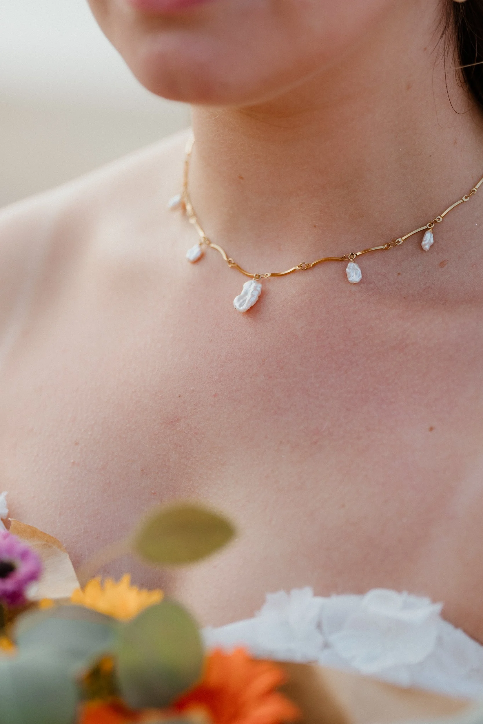 Close-up of a woman's neck wearing a gold necklace with white pearl-like pendants, with a blurred floral arrangement in the foreground.