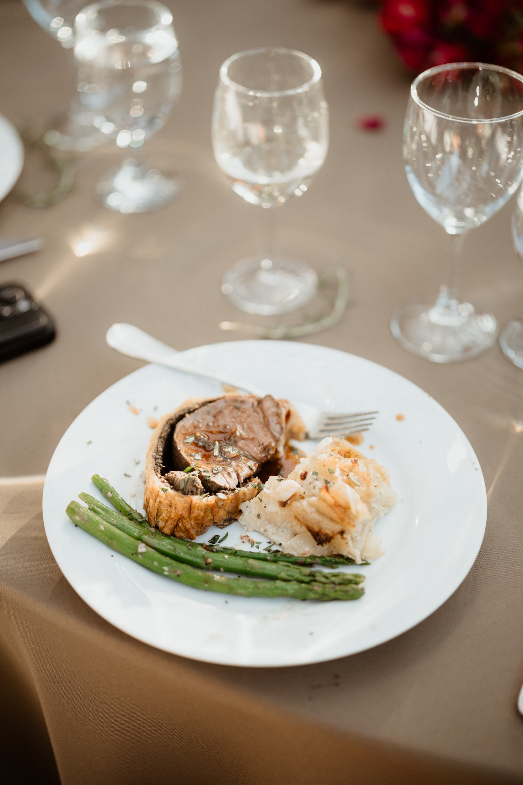 A dinner plate with a serving of beef Wellington, mashed potatoes, and green asparagus on a dining table with empty wine glasses in the background.