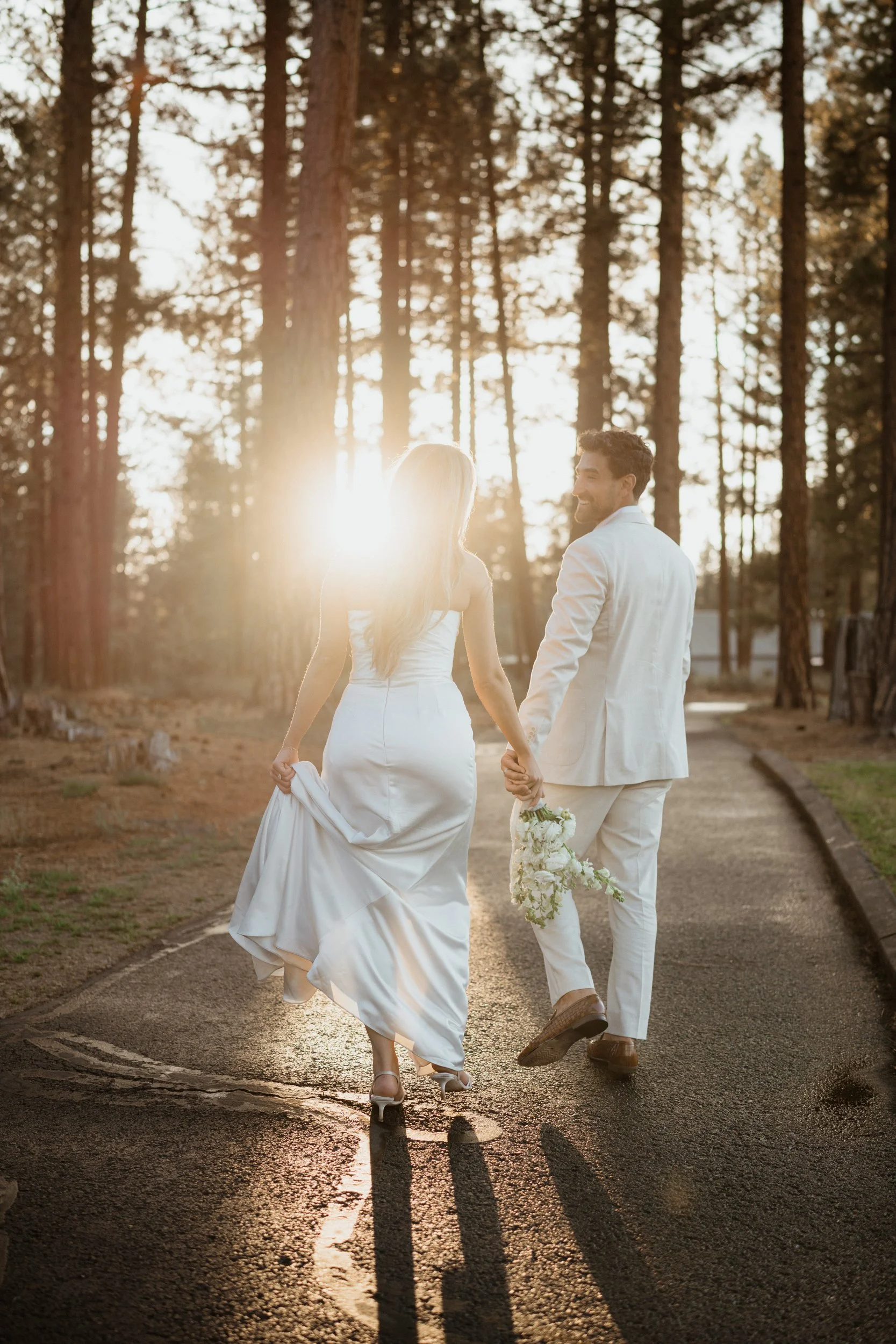 A newlywed couple holding hands and walking down a forest path at sunset, with the bride holding a bouquet and wearing a white wedding dress, and the groom in a light-colored suit.