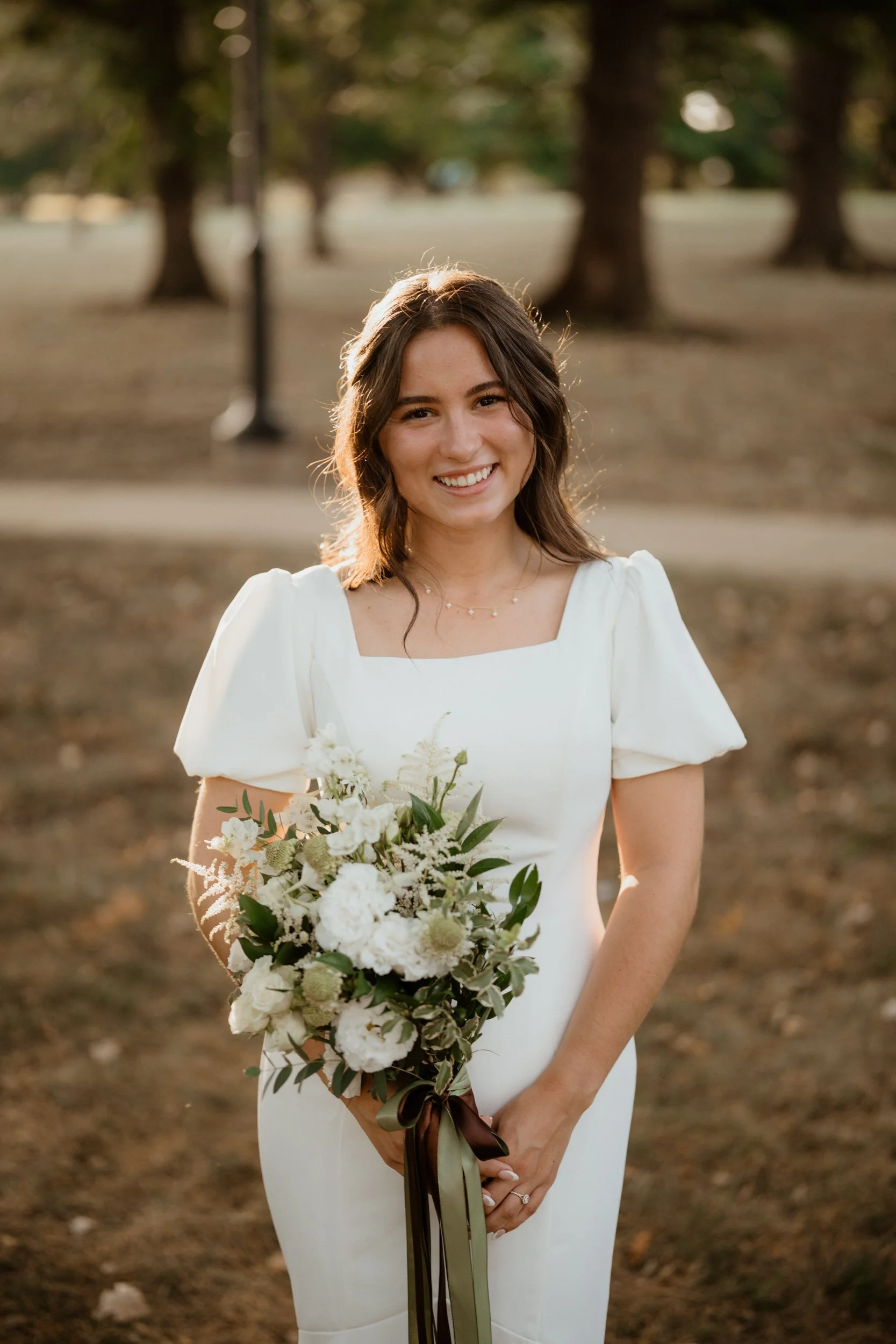 A smiling young woman in a white dress holding a bouquet of white and green flowers, standing outdoors in a park with trees in the background during sunset.