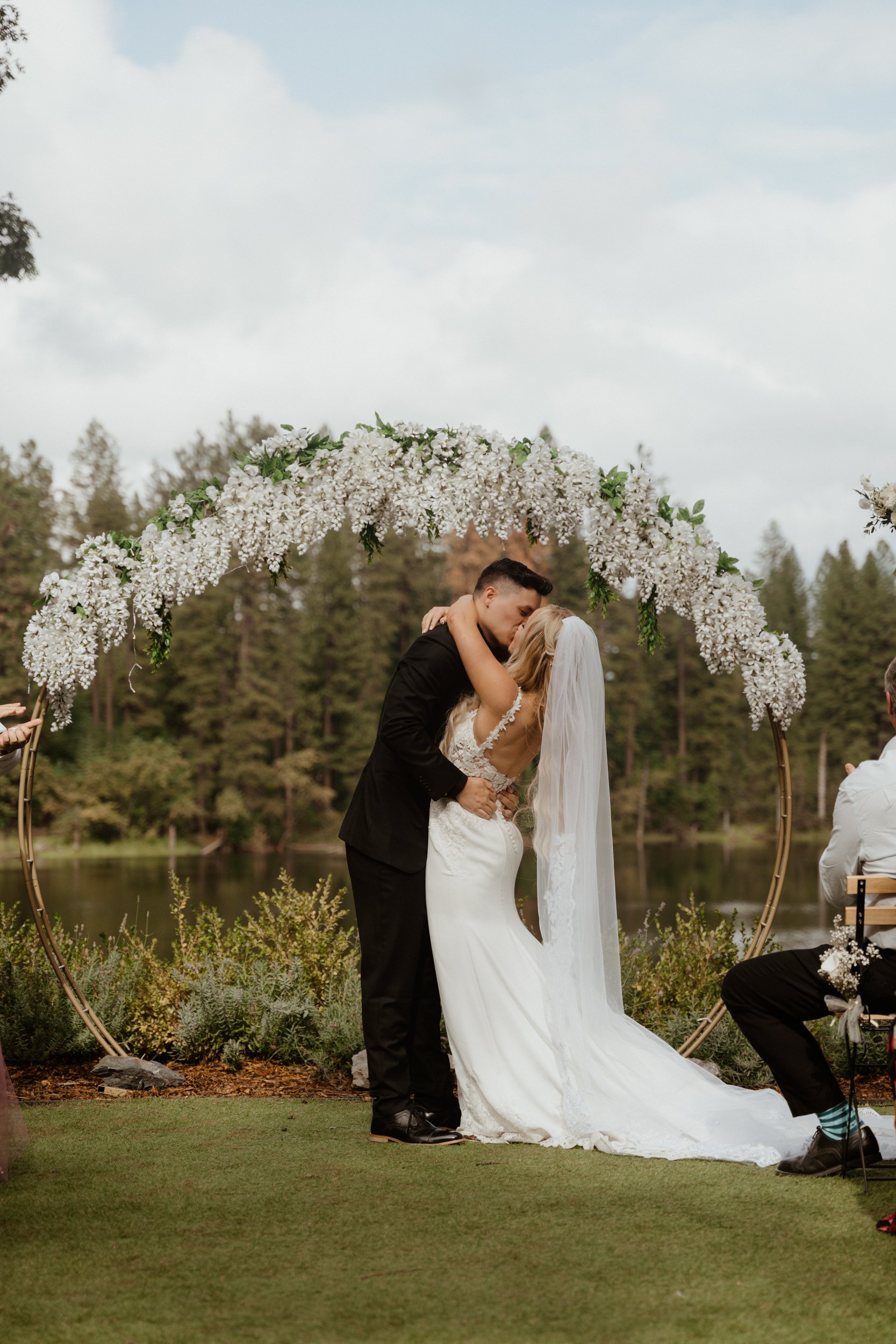 A newlywed couple hugging and kissing under a floral arch during an outdoor wedding ceremony by a lake with trees in the background.