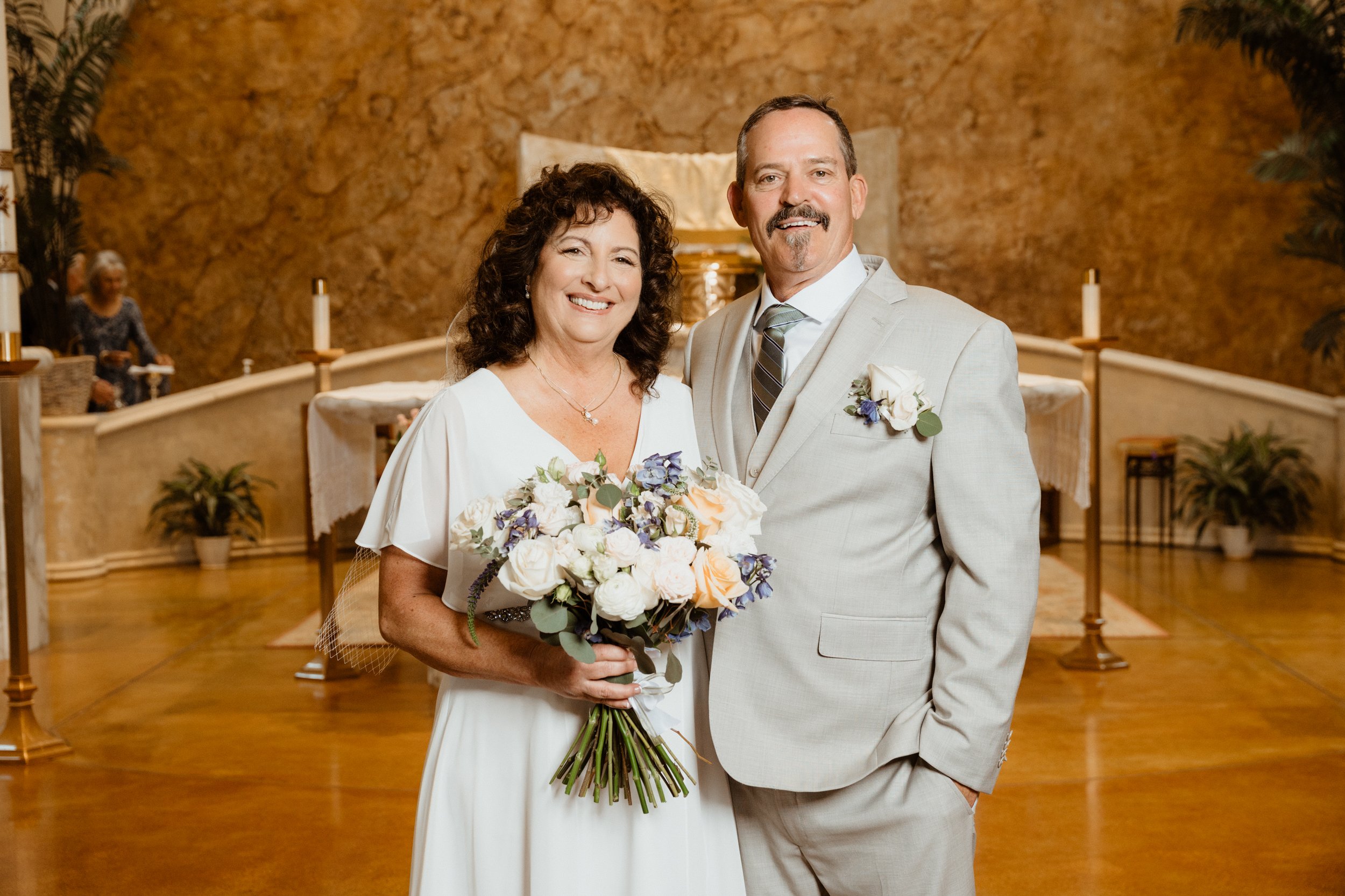 A bride and groom smiling, standing inside a church with a rustic brick background, the bride holding a bouquet of white and purple flowers and wearing a white dress, the groom in a light gray suit with a white shirt and tie, both with boutonnieres, 