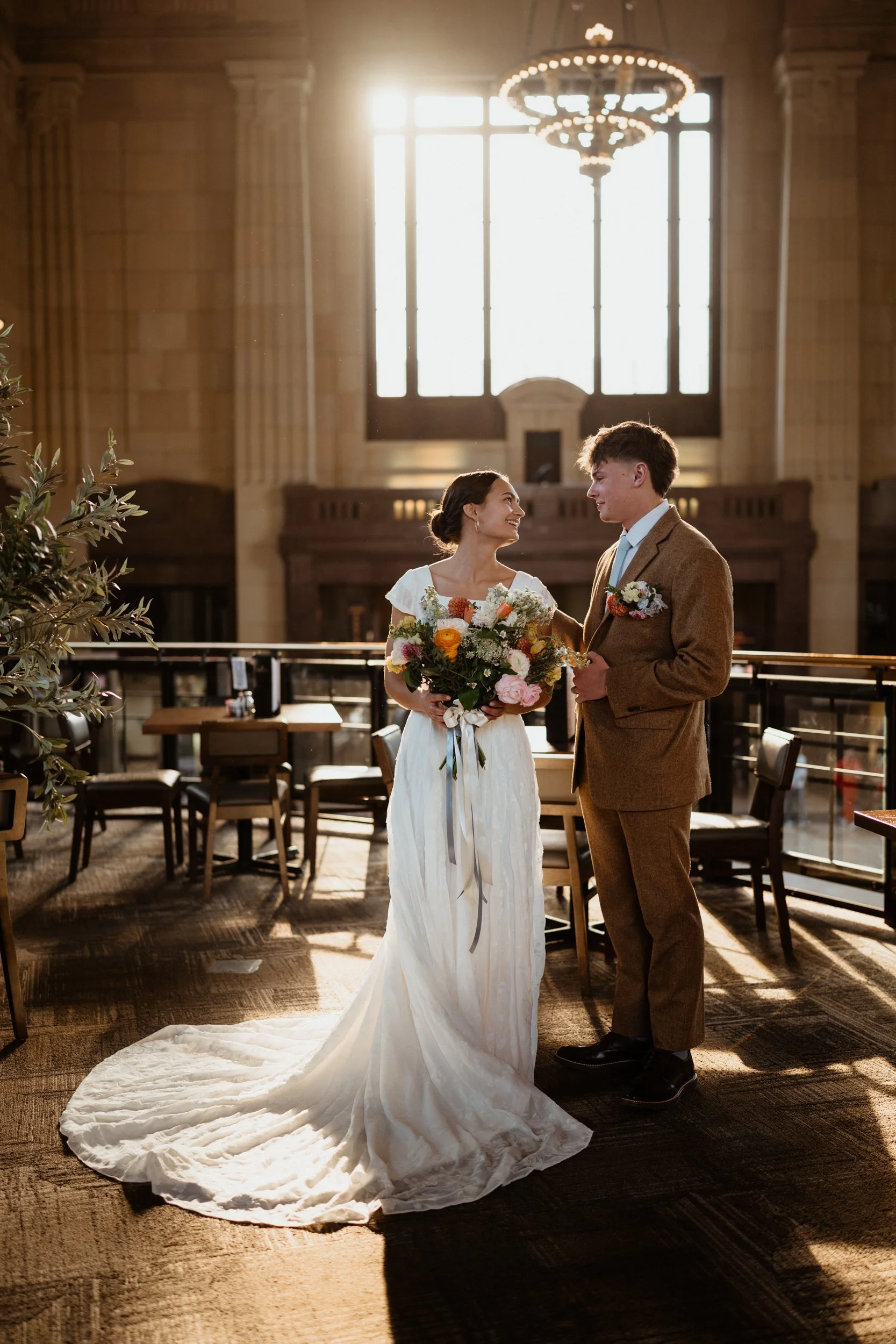A bride and groom stand facing each other in a warmly lit indoor venue, with large windows and a chandelier overhead. The bride holds a colorful bouquet and wears a white wedding dress with a long train; the groom wears a brown suit. They smile and look at each other affectionately.