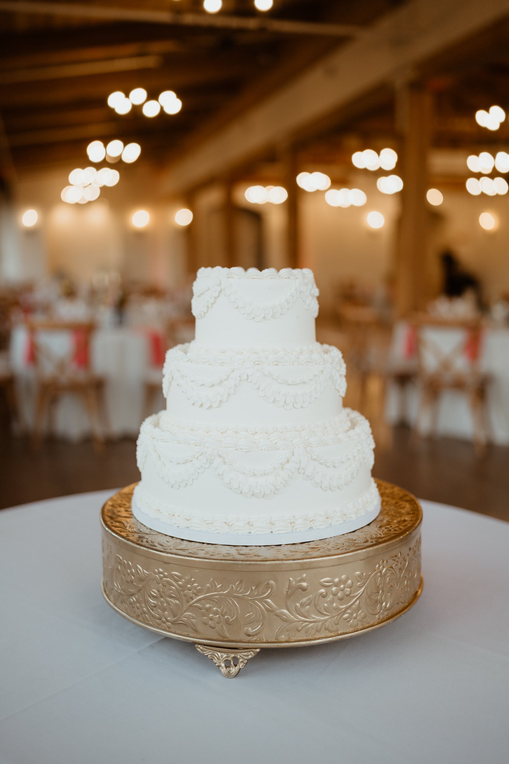 A three-tiered white wedding cake with piped decorative icing, placed on a gold ornate cake stand in a decorated reception hall.