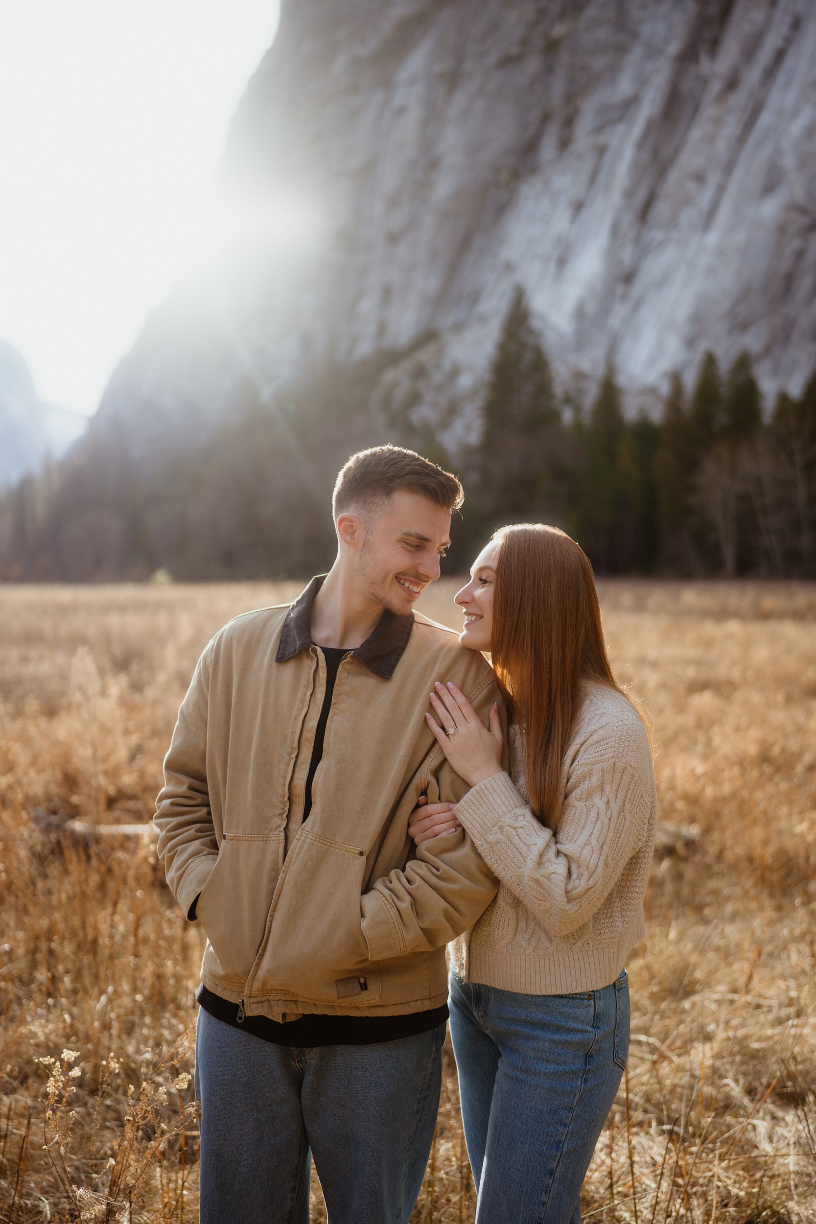 A smiling young couple in casual clothes stands close together in a field with tall grass, with mountains and trees in the background, illuminated by sunlight.