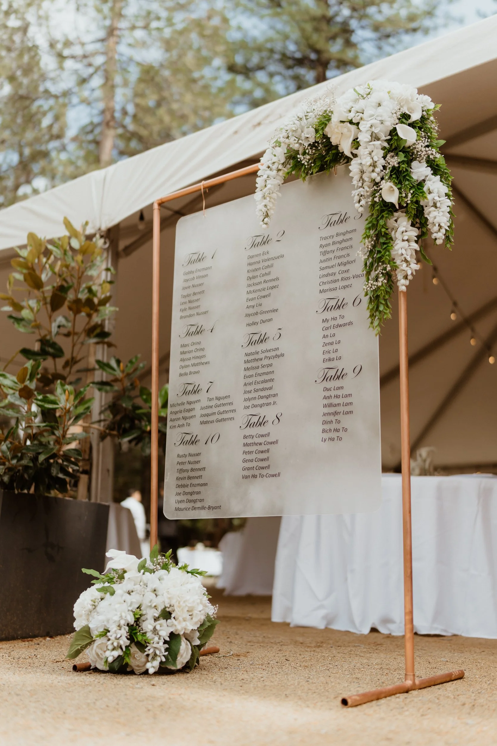 Wedding seating chart presented on a white board with floral decorations, placed outdoors near a white tent.