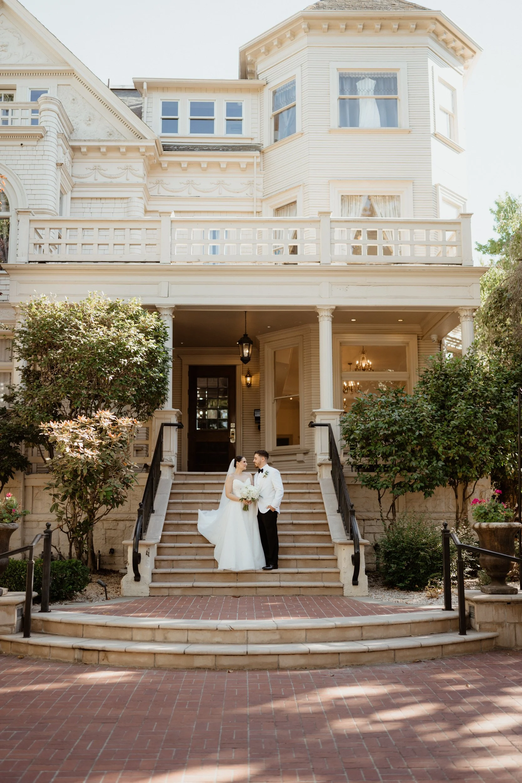 Bride and groom standing on the steps of a large, vintage, white house with trees and shrubs around.