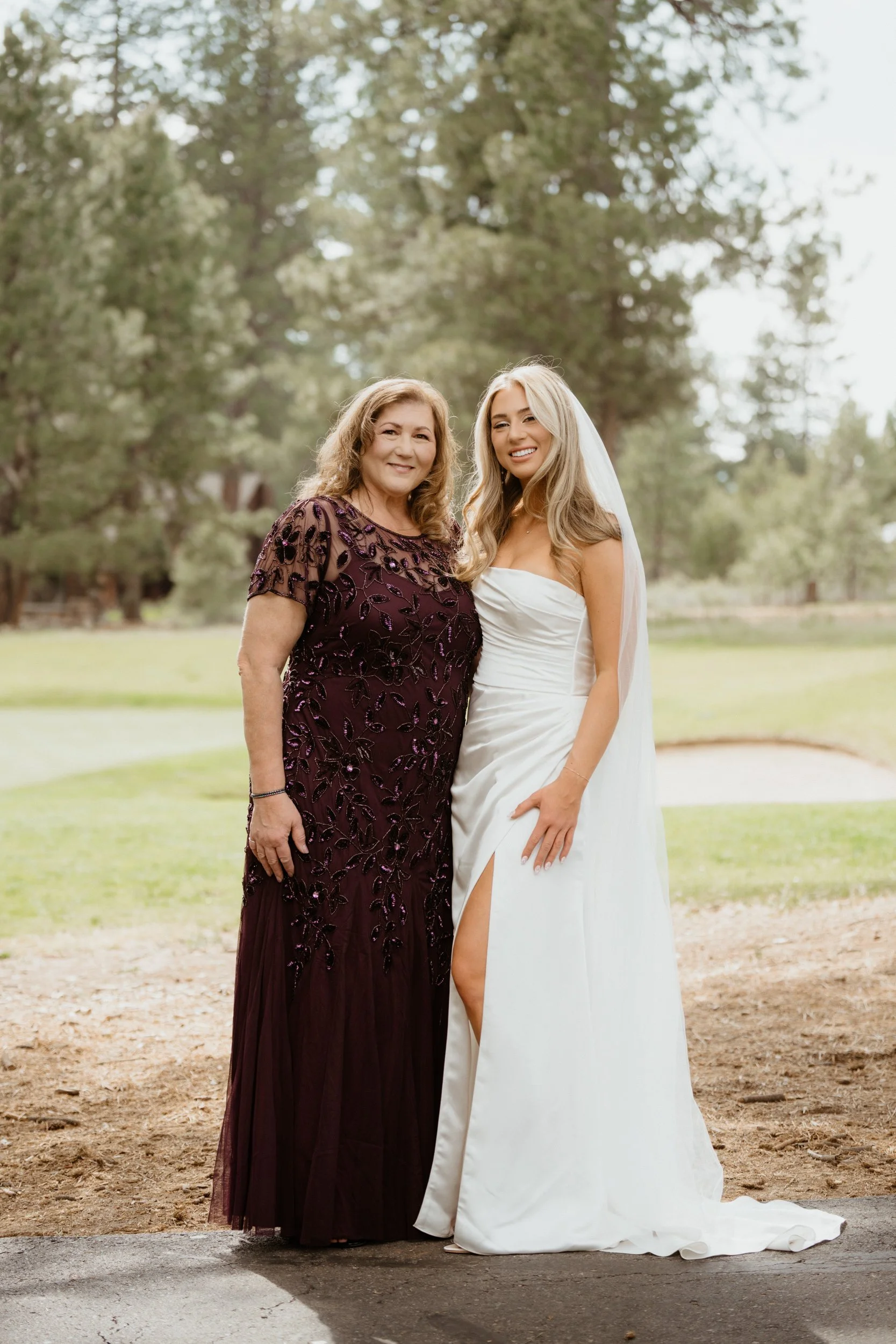 A woman in a dark purple, embellished dress and a bride in a white strapless gown and veil stand together outdoors on a lawn with trees in the background, smiling at the camera.