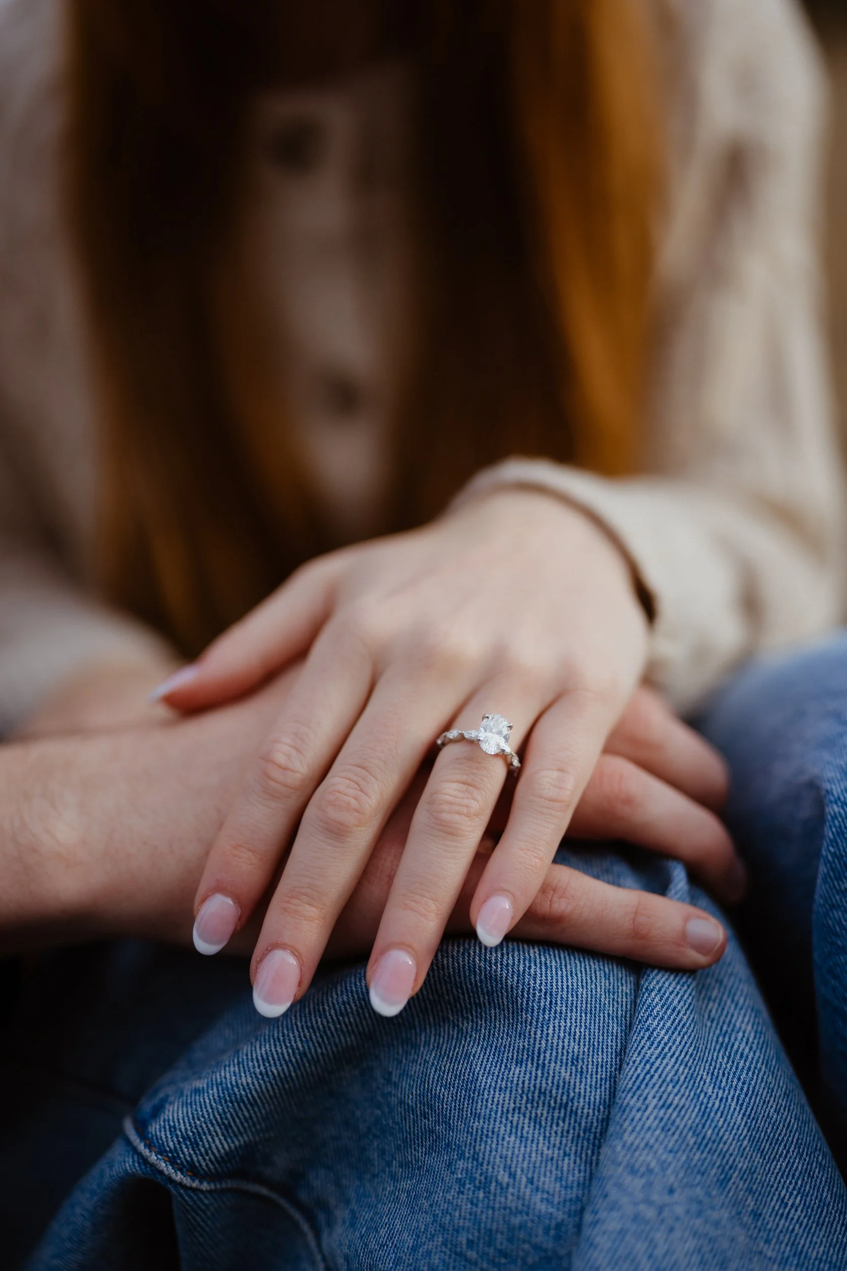 Close-up of a woman’s hand with a large engagement ring resting on her friend's knee, wearing jeans and a beige top, with a blurred background.