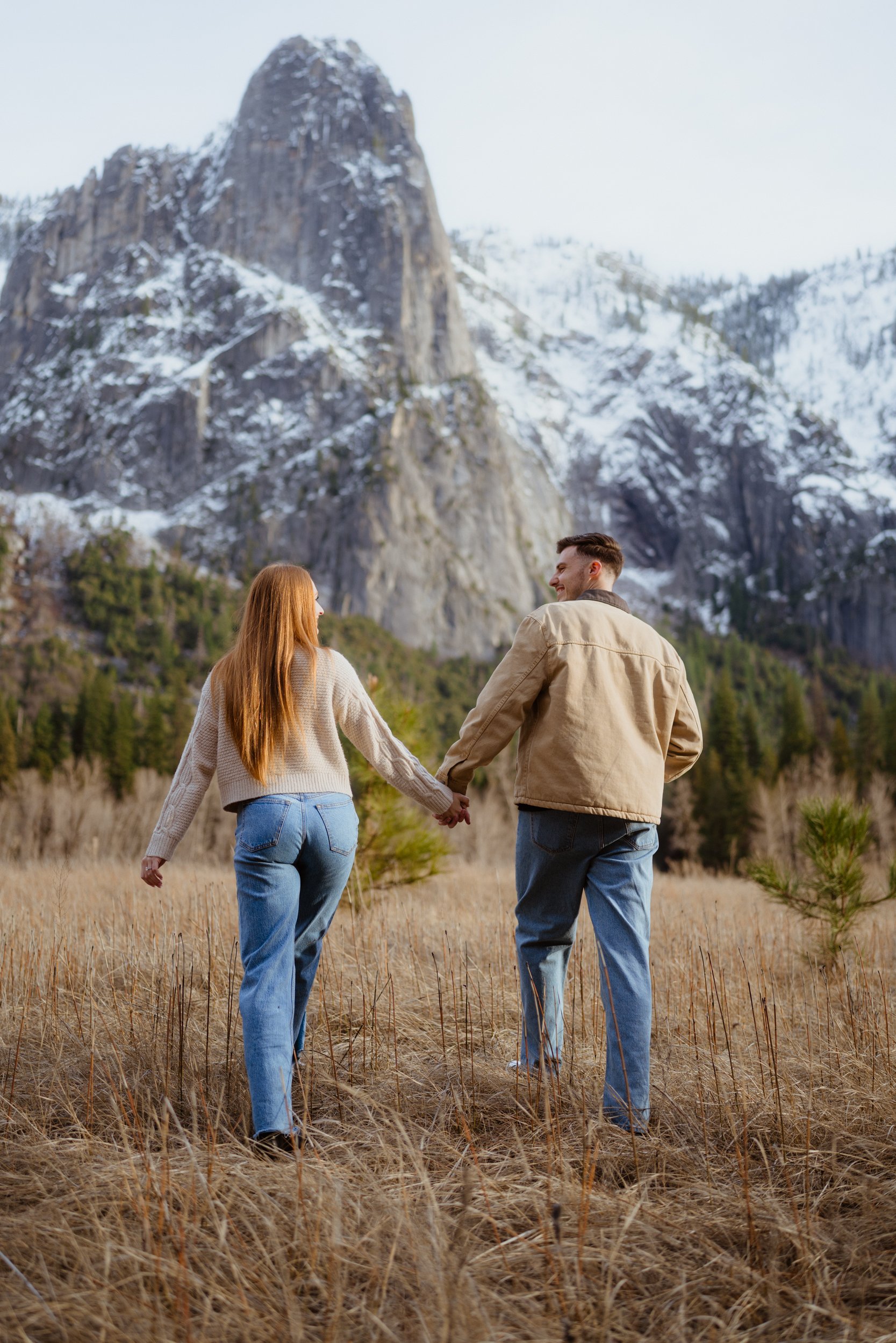 A couple holding hands walking through a grassy field with mountains in the background.