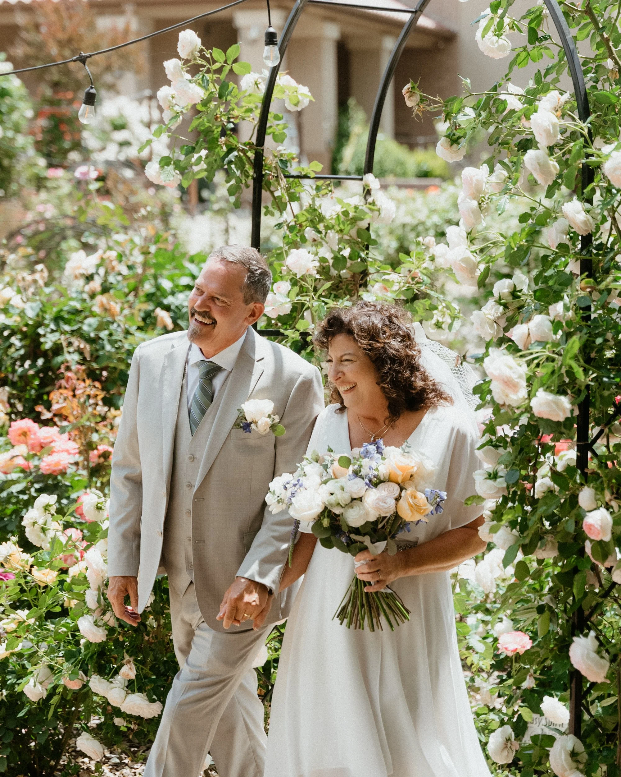 A bride and groom walking arm in arm under a floral arch, smiling, with the bride holding a bouquet of flowers. The scene is outdoors with flowers and greenery around.