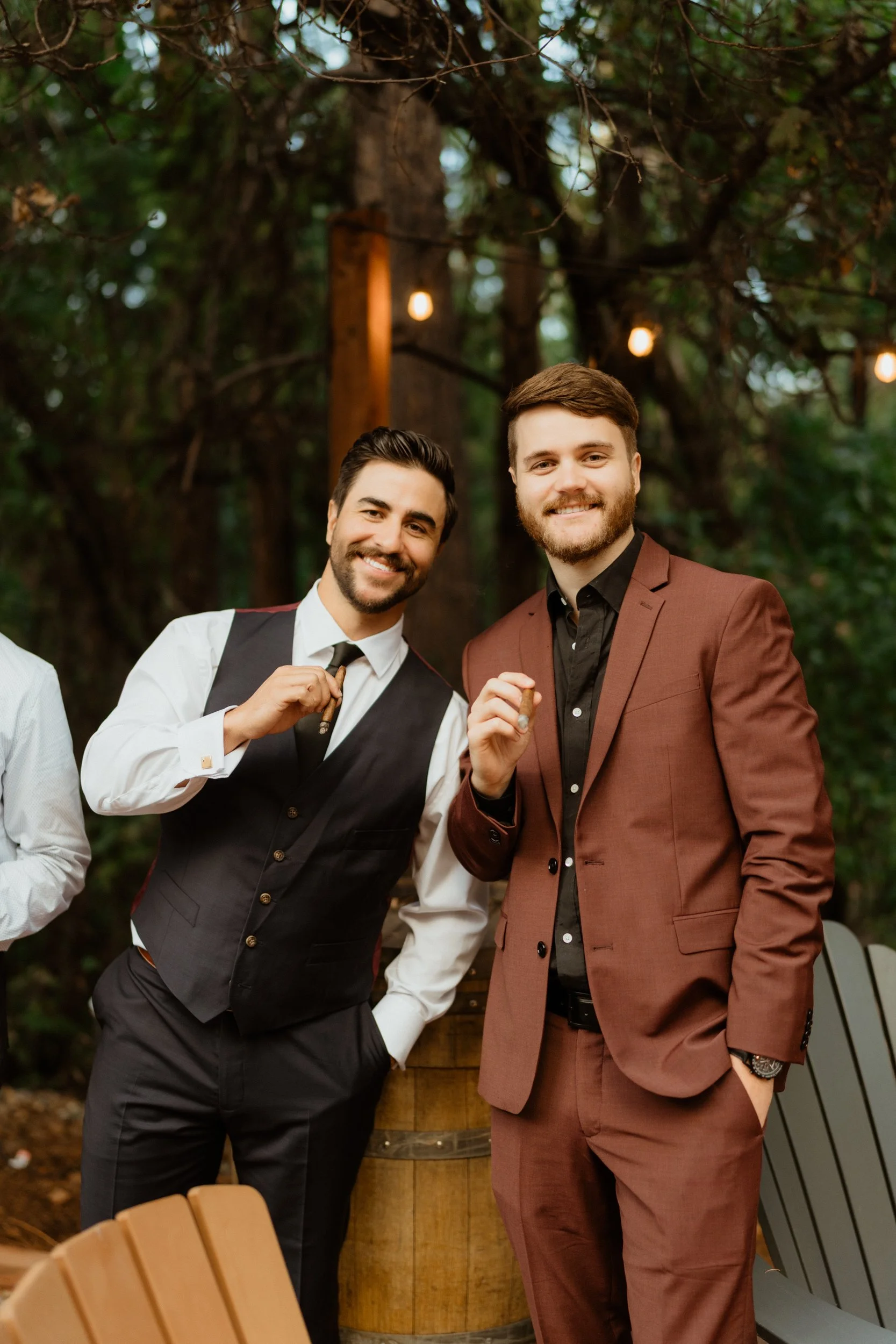 Two smiling men in suits holding cigars at an outdoor gathering with trees and string lights in the background.