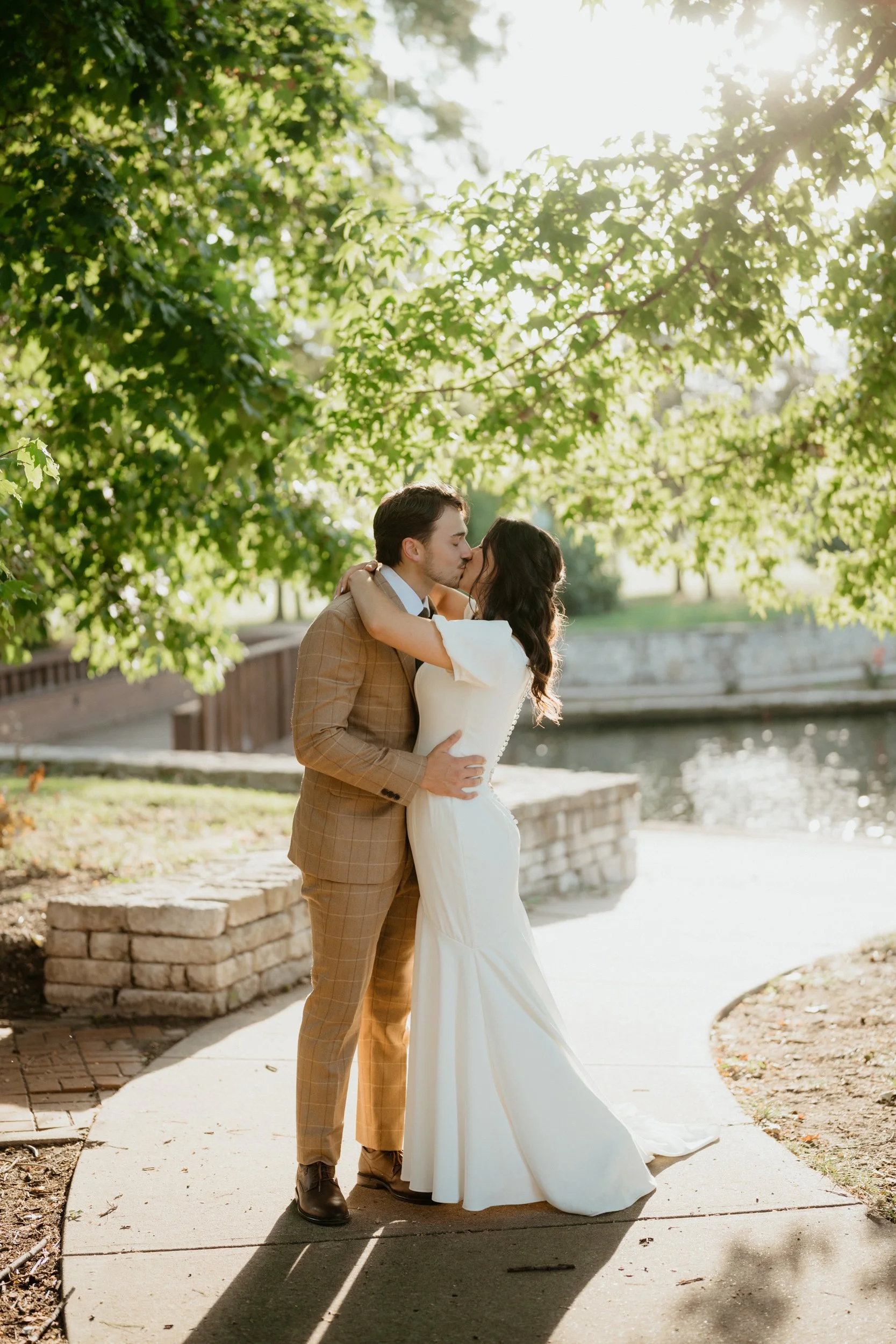 A couple dressed in wedding attire sharing a kiss outdoors near a river, with sunlight filtering through green trees.