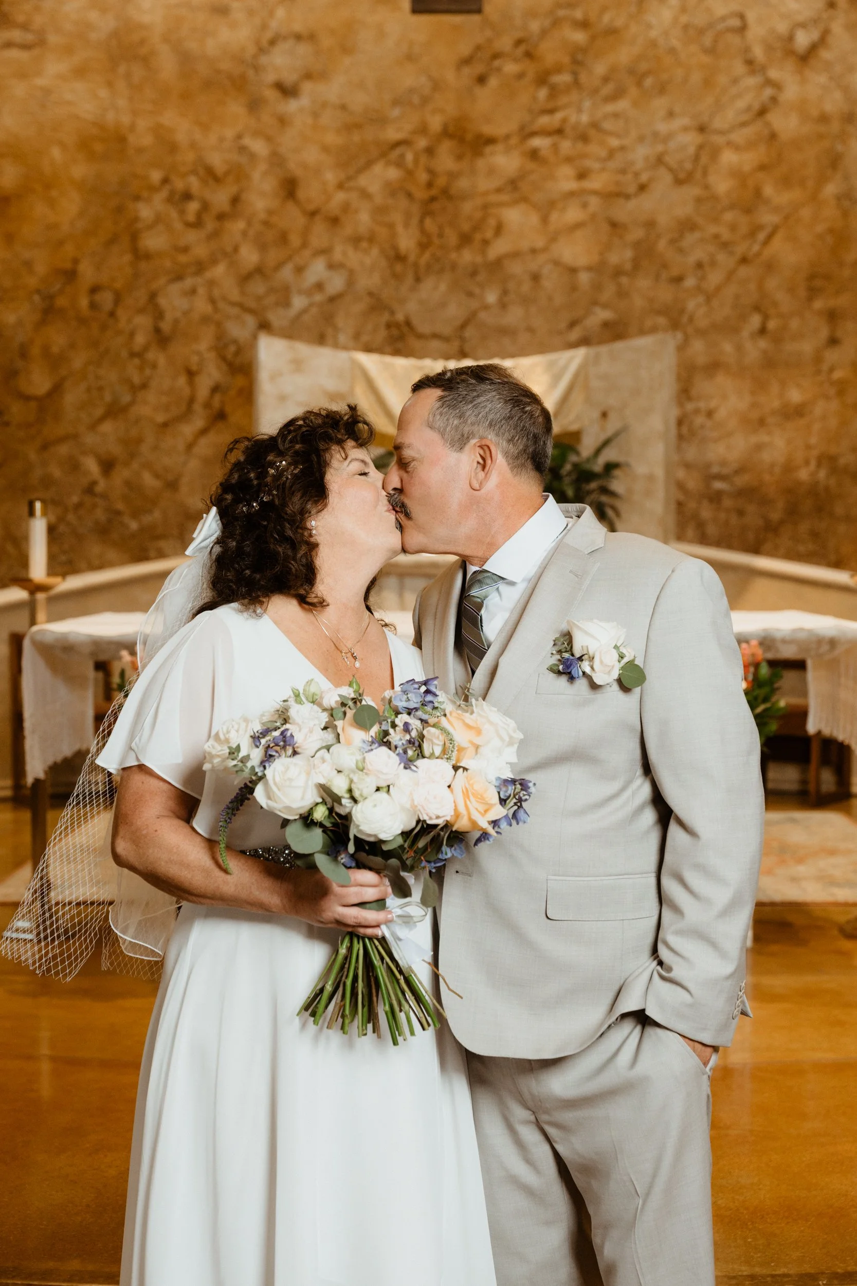 A bride and groom sharing a kiss during their wedding ceremony inside a church, with the bride holding a bouquet of flowers.