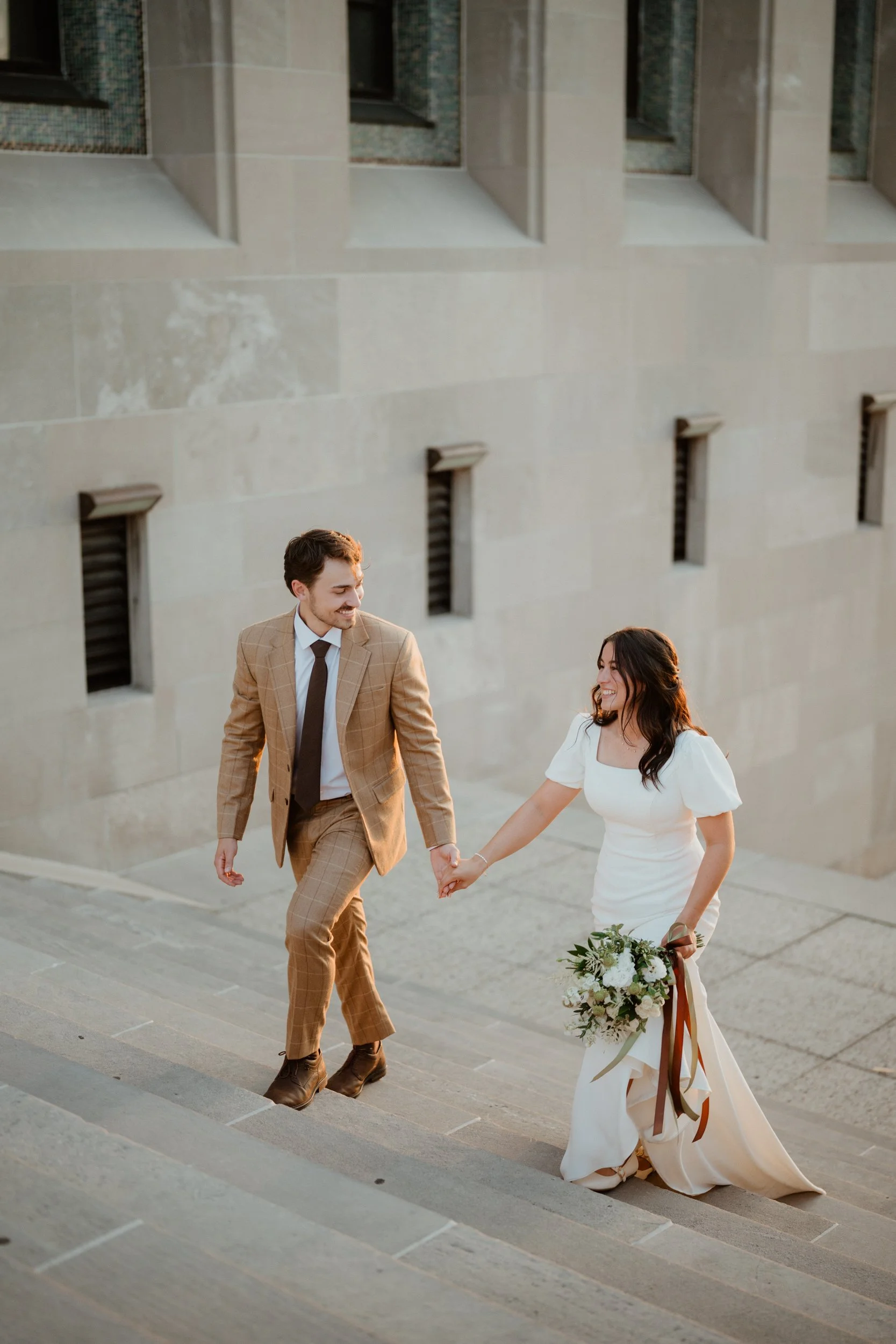 A happy couple walking up outdoor stone steps, holding hands, with the woman holding a bouquet of flowers, during sunset.