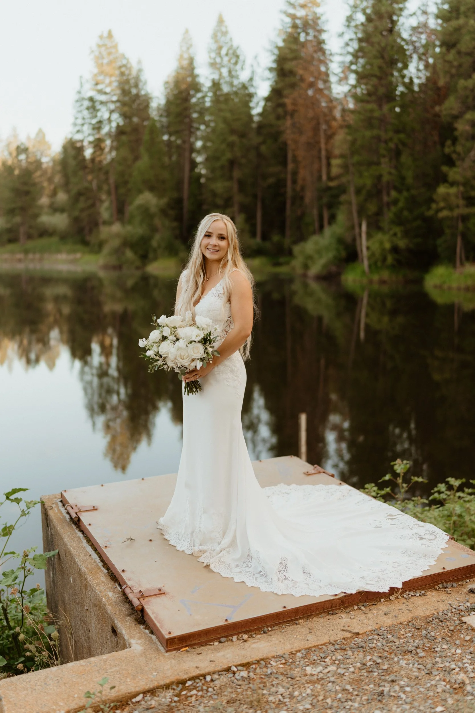 A smiling bride in a white lace wedding gown holding a bouquet of white flowers, standing on a dock by a calm lake surrounded by green trees.