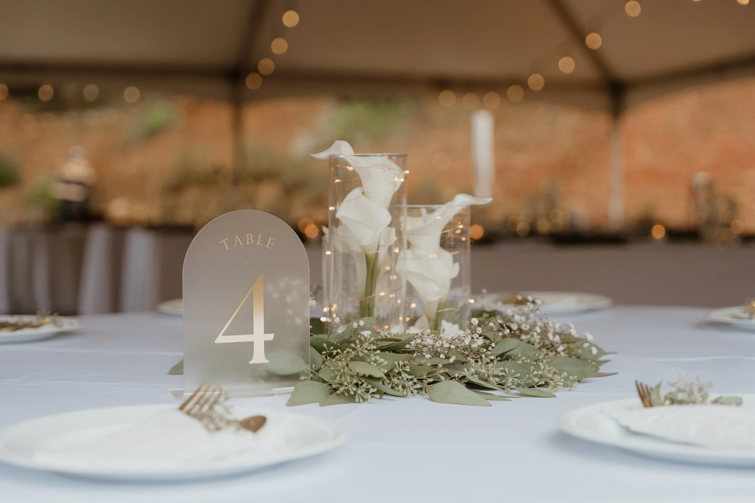 Wedding reception table with white plates, a frosted acrylic table number sign displaying number 4, floral centerpiece with white calla lilies, greenery, and small white flowers, under a tent with string lights, and a blurred brick wall background.