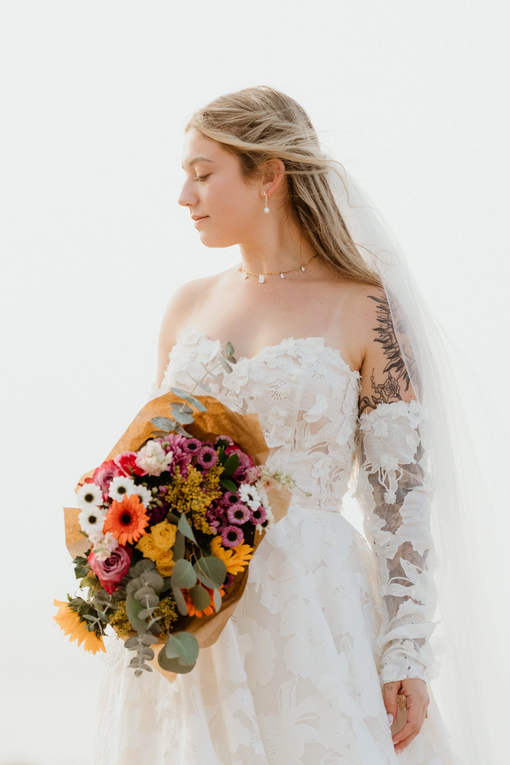 A bride with long blonde hair holding a colorful bouquet of flowers, wearing a white lace off-shoulder wedding dress with sheer lace sleeves and jewelry, standing against a plain white background.