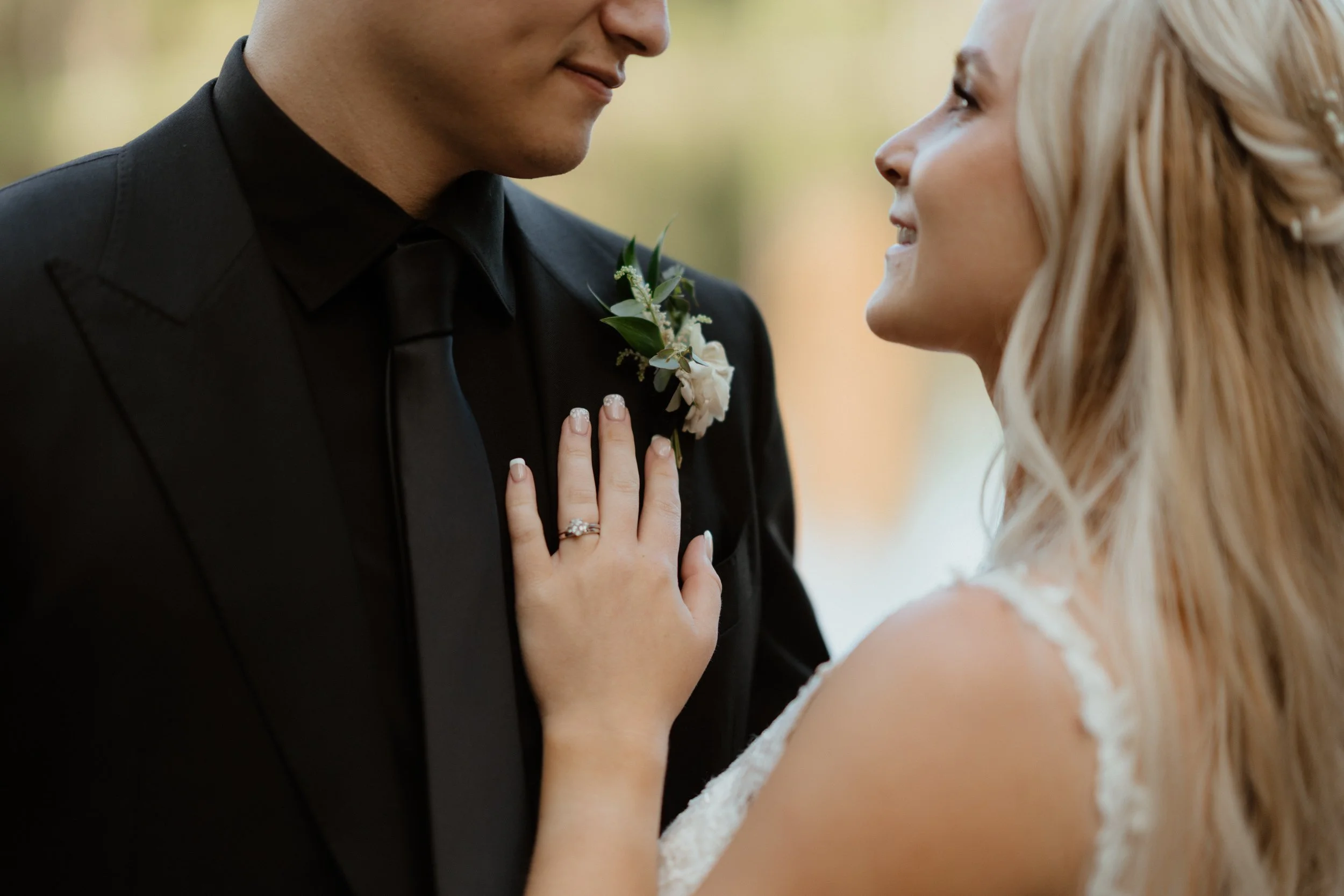 A bride and groom facing each other, the bride touching the groom's chest with her hand showing her wedding ring, both smiling.