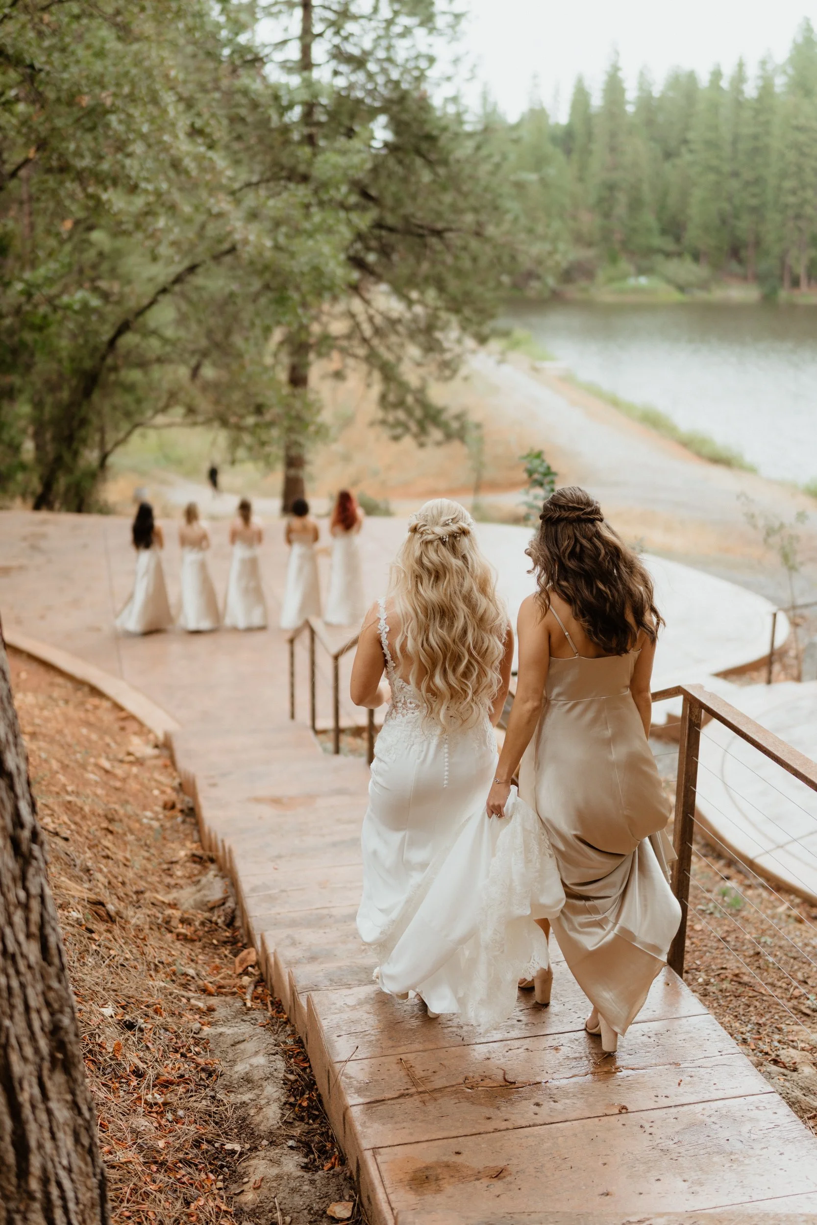 Two women in champagne-colored dresses walking down a wooden path towards six women in white dresses by a lake with trees in the background.