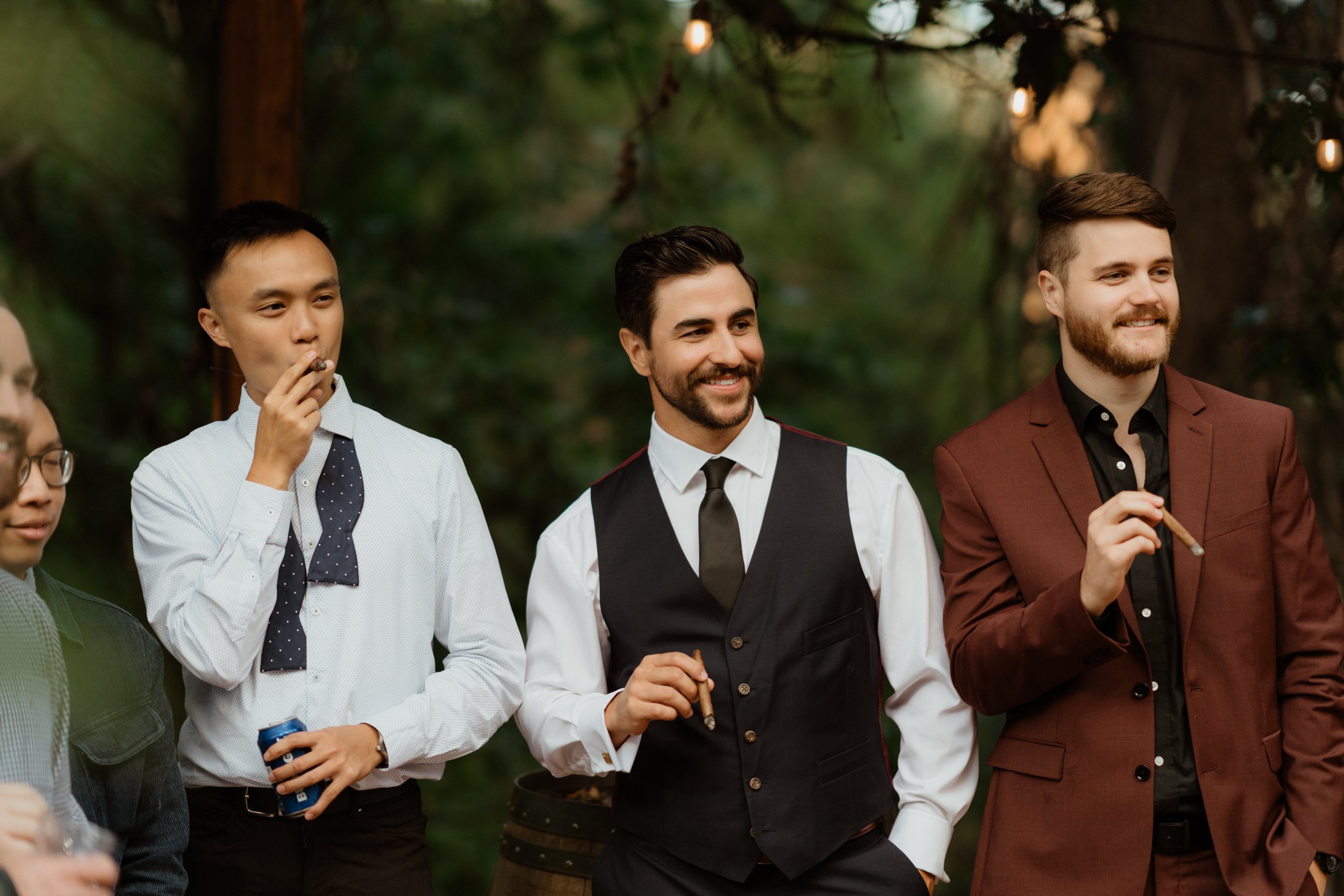 Group of men in formal and semi-formal attire enjoying outdoor party, some smoking cigars, standing among trees with string lights overhead.