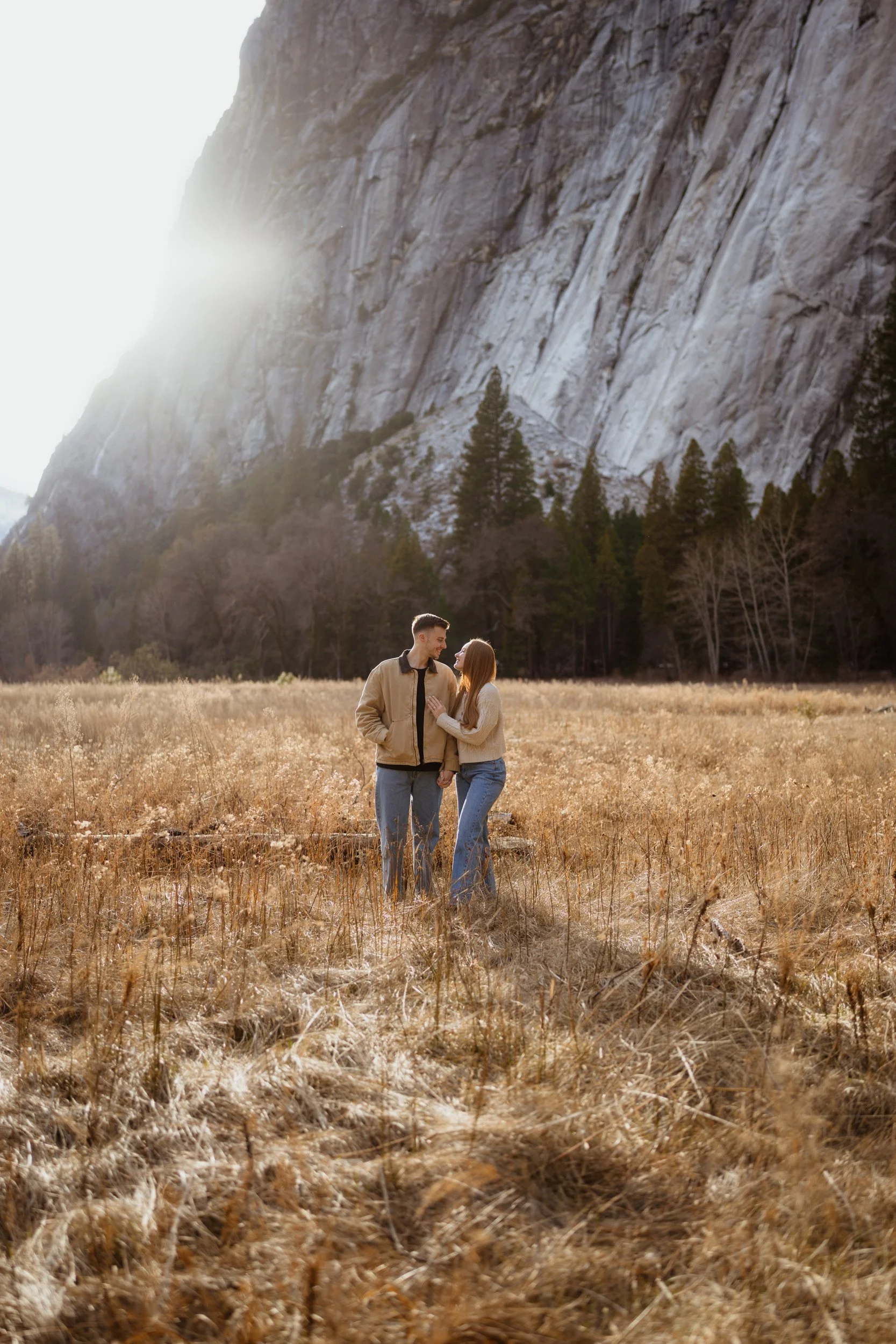 A couple standing close together in a grassy field with mountains and trees in the background during sunset.