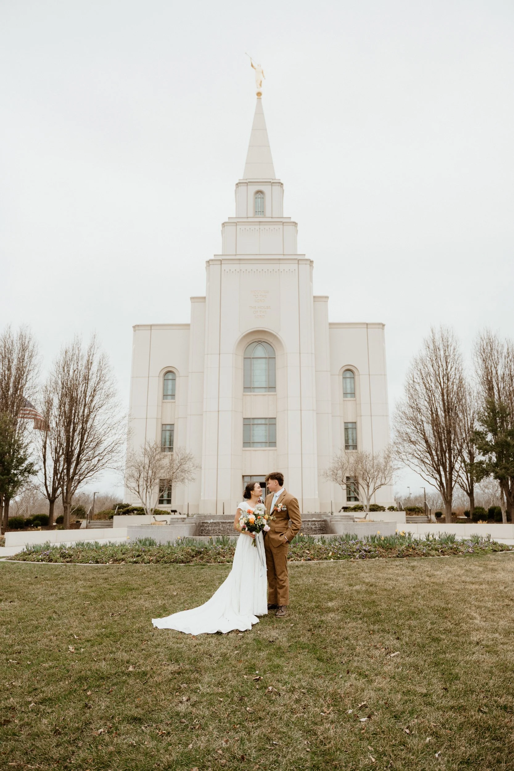 A bride and groom standing close together in front of a white church, looking at each other. The bride is holding a bouquet of flowers, and the groom is dressed in a brown suit. The church has a tall steeple and is surrounded by leafless trees and a 