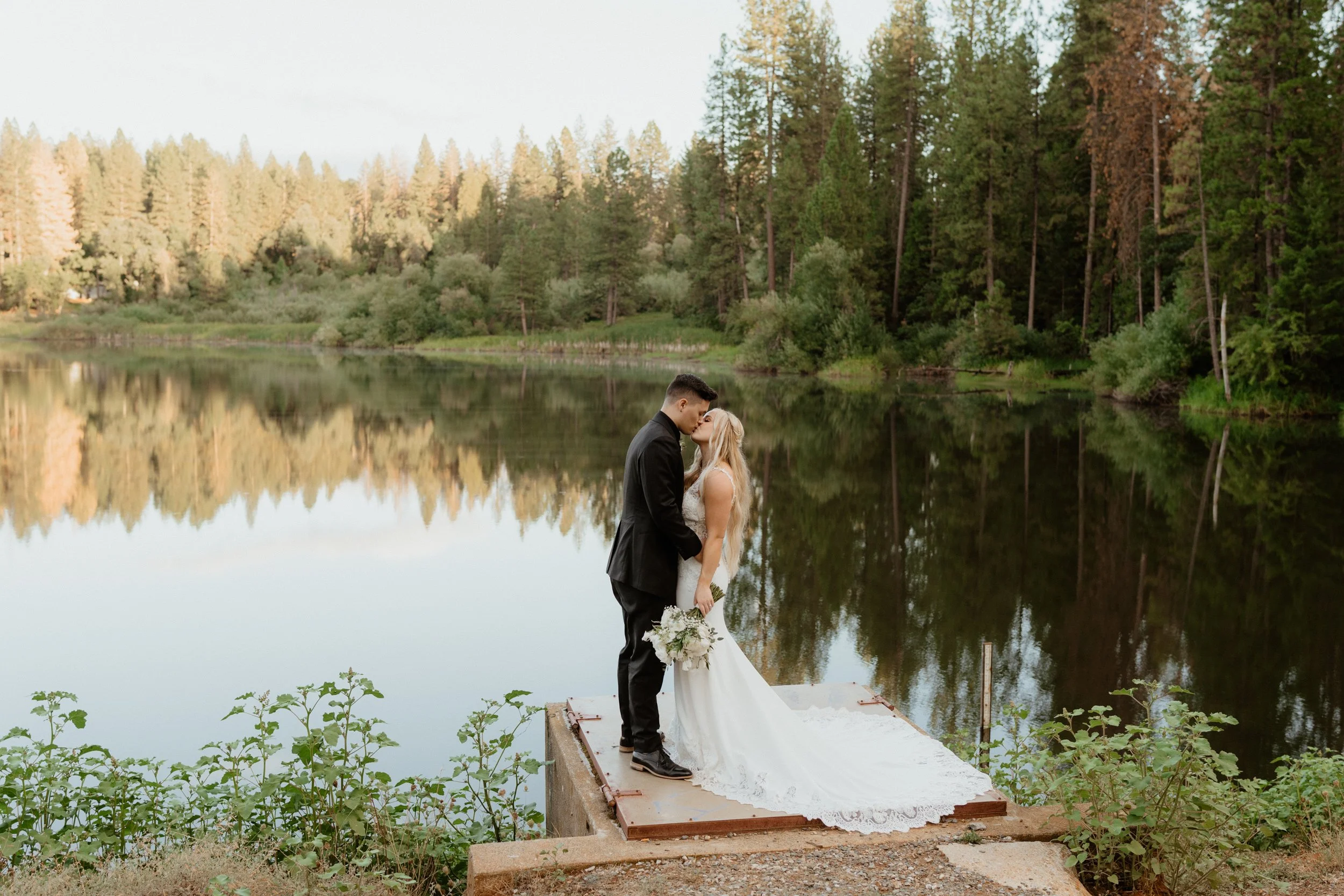 A newlywed couple sharing a kiss on a dock by a lake surrounded by trees.