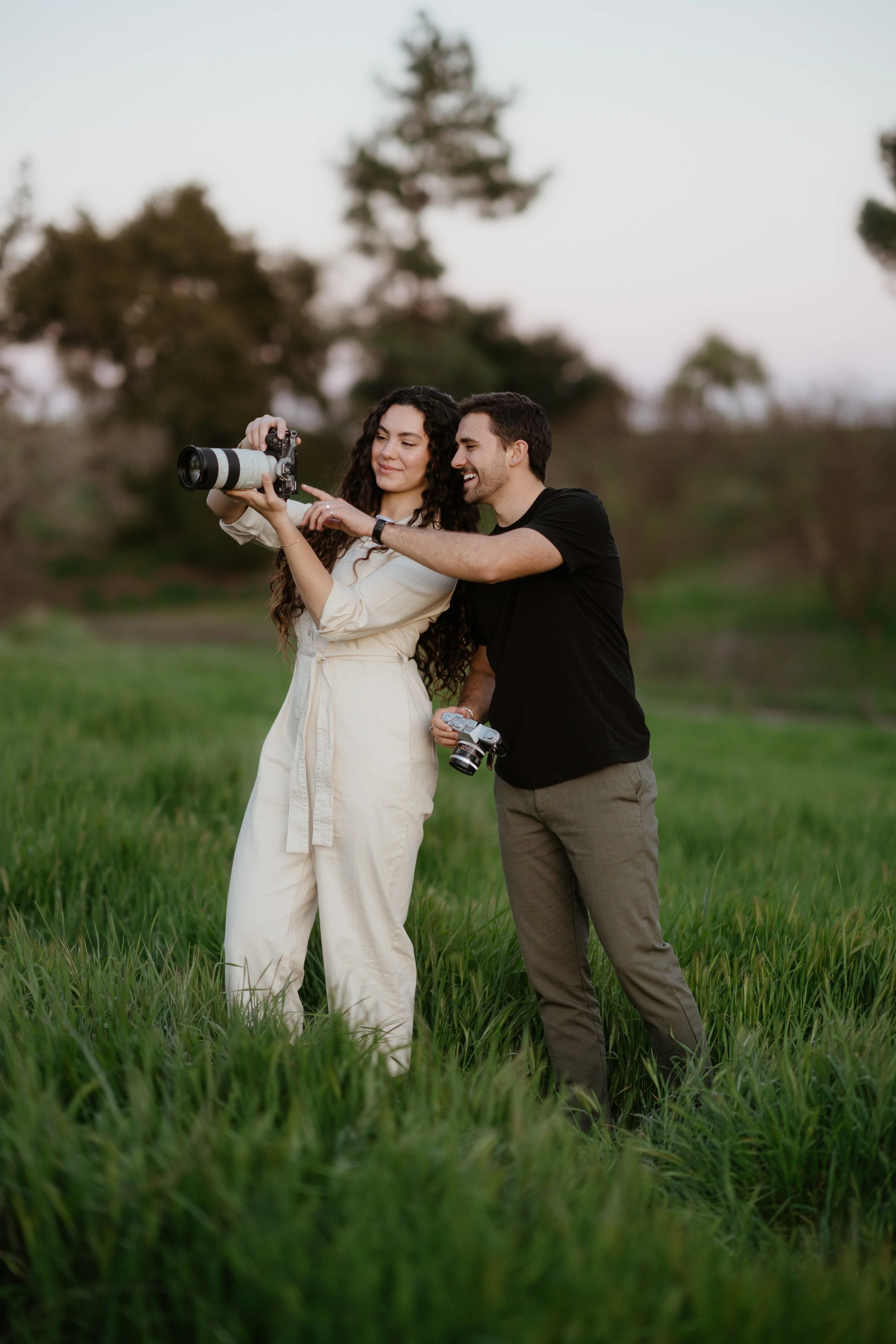 A man and a woman standing in a grassy field, both holding cameras, with the man showing the woman something on the camera's screen, in an outdoor setting during early evening.