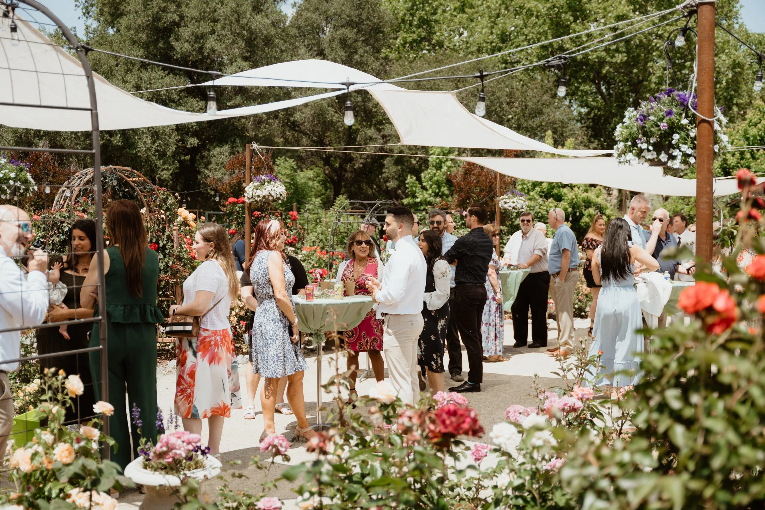 People gathered outdoors at a garden party with floral decorations and shaded areas, socializing and enjoying the event.