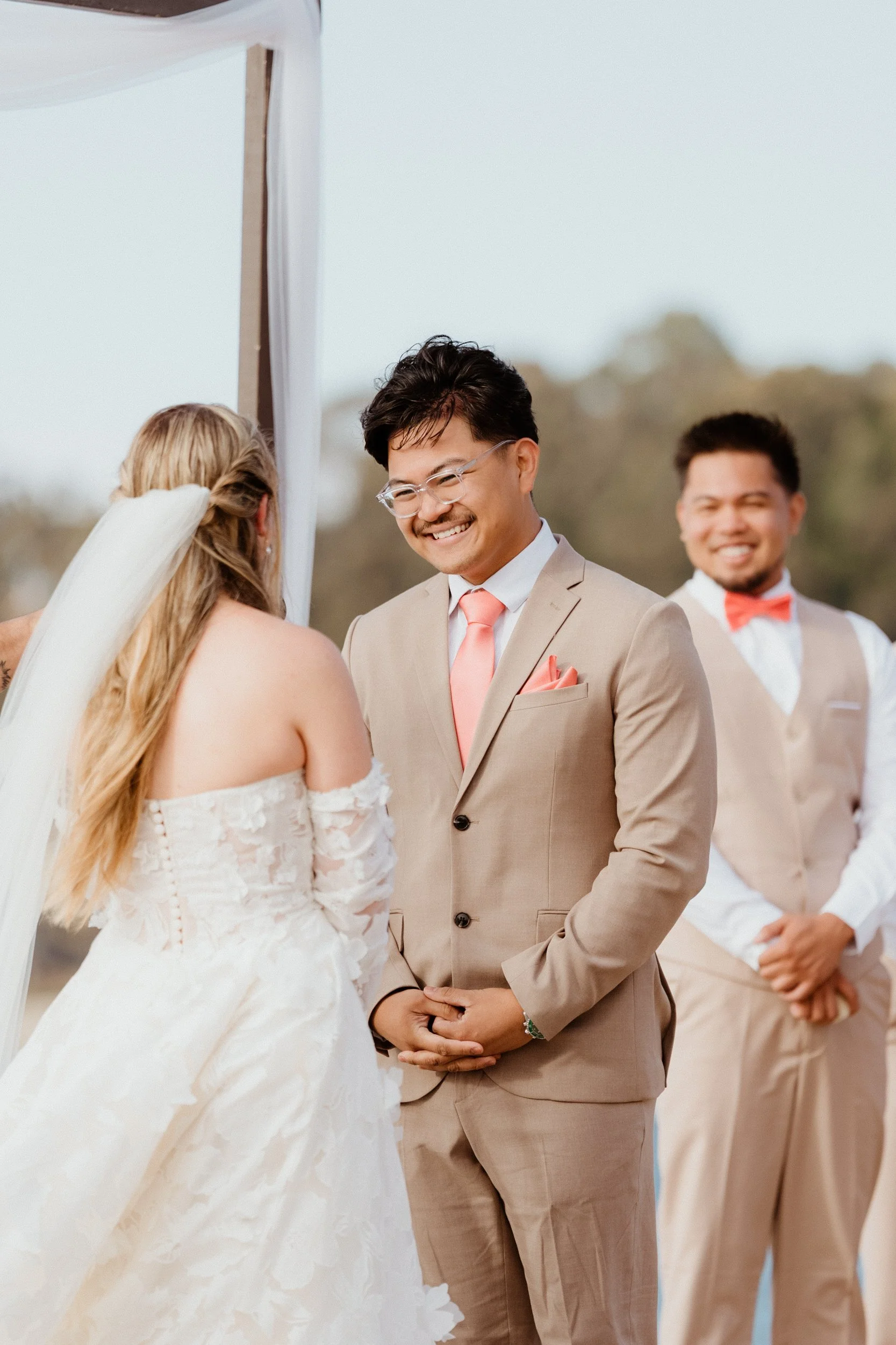 A wedding ceremony with a couple exchanging vows, officiant smiling in background, outdoor setting.