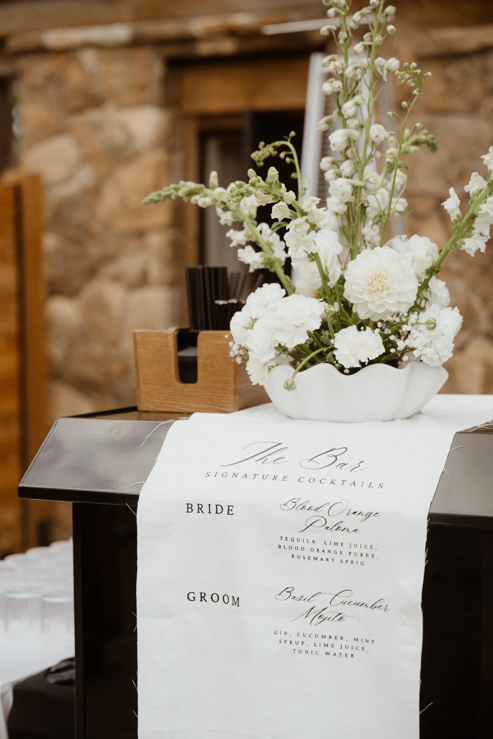 A floral arrangement of white flowers in a white vase on a table with a white tablecloth, with a menu listing signature cocktails for a wedding.
