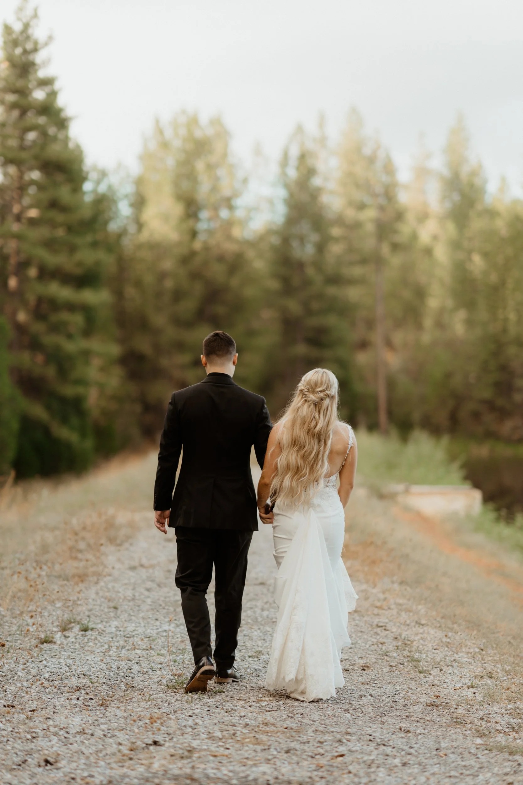 A newlywed couple walks hand in hand down a gravel path through a forest, with the bride in a white wedding dress and the groom in a black suit.