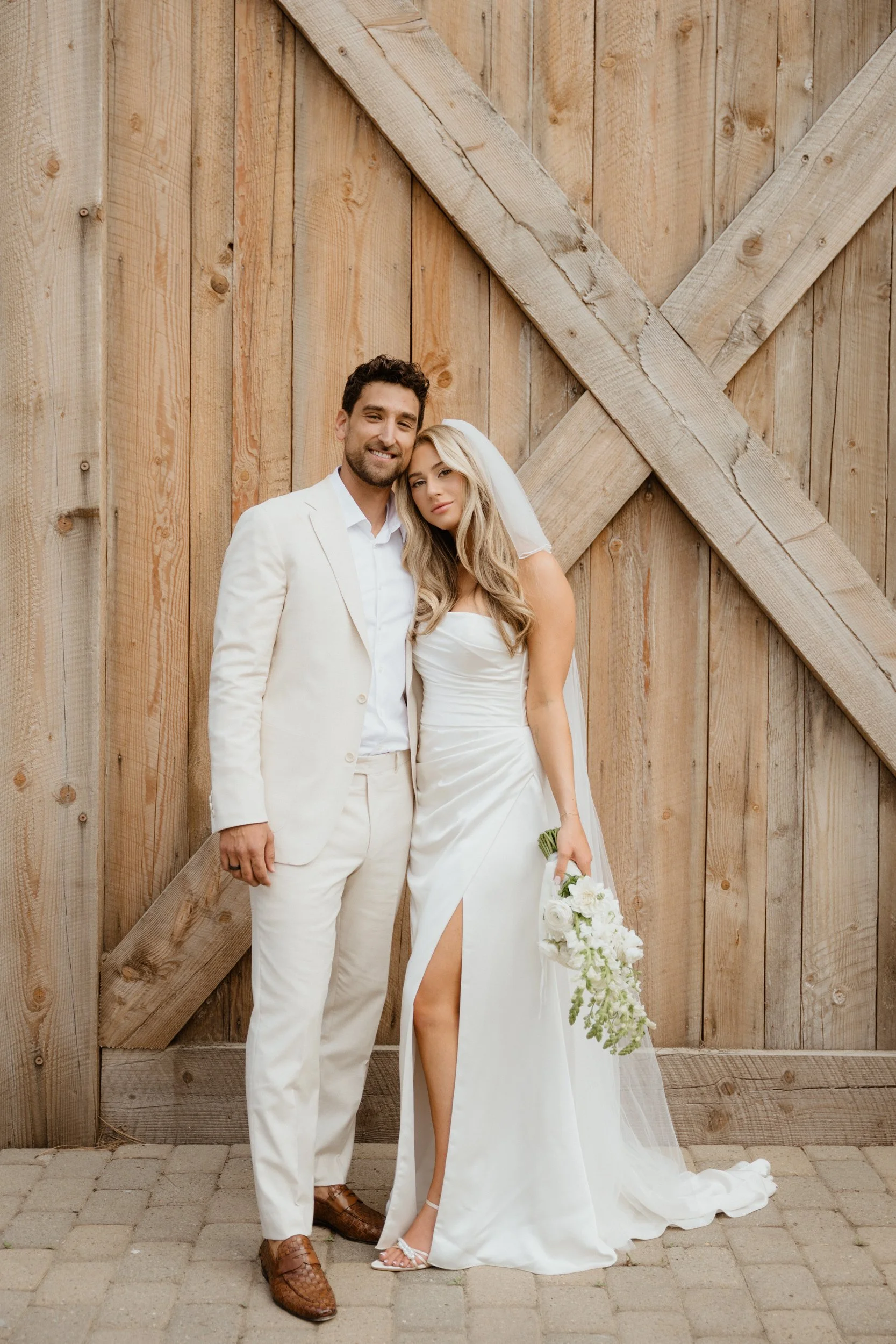 A bride and groom standing close together in front of a large wooden barn door, smiling for a photo. The groom is wearing a white suit, and the bride is in a white wedding dress with a high slit, holding a bouquet of white flowers.