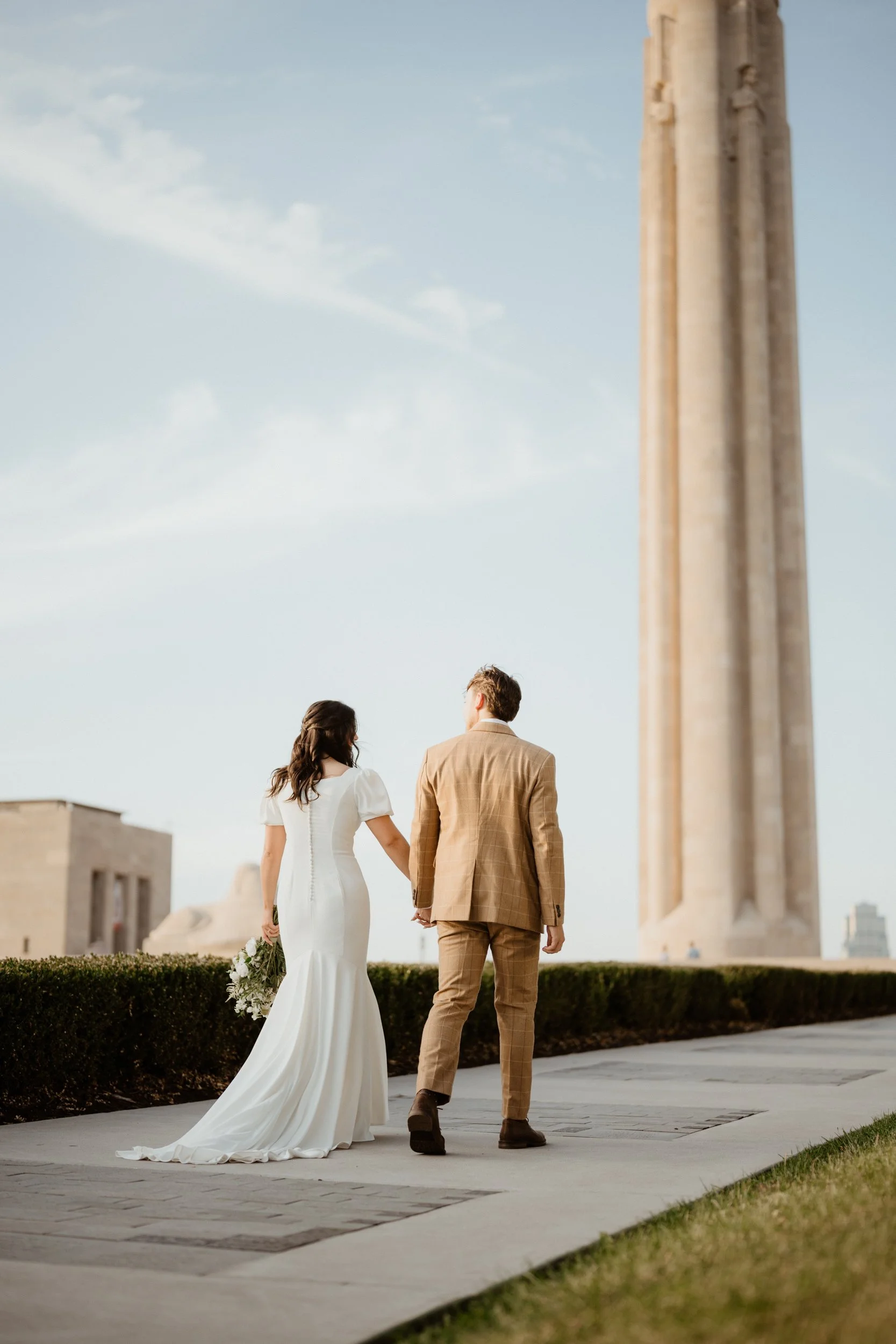 A bride and groom walking hand in hand outdoors near a tall historical monument, with clear blue sky above.