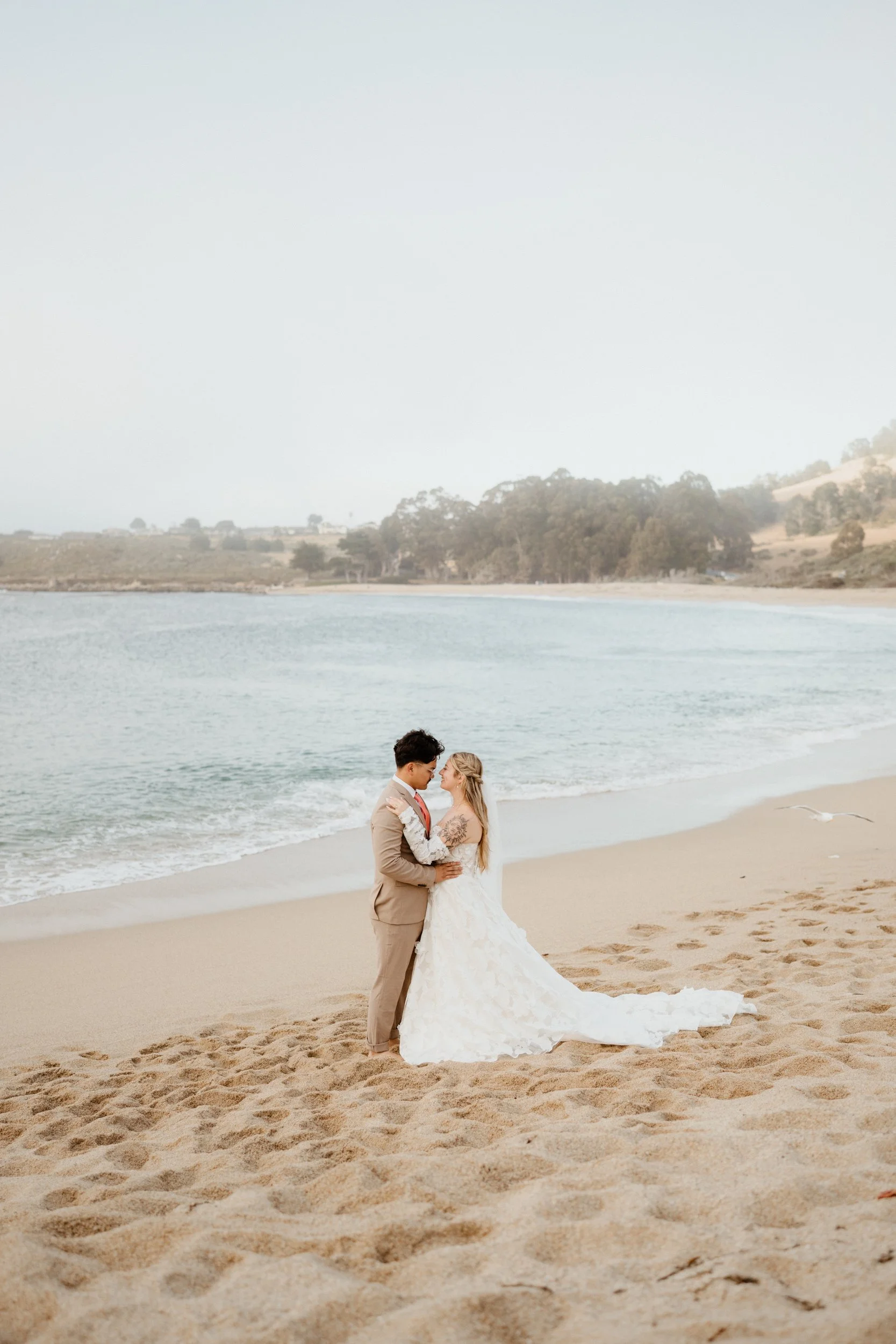 A bride and groom standing close on a sandy beach, gazing into each other's eyes, with the ocean behind them and a distant hilly shoreline.