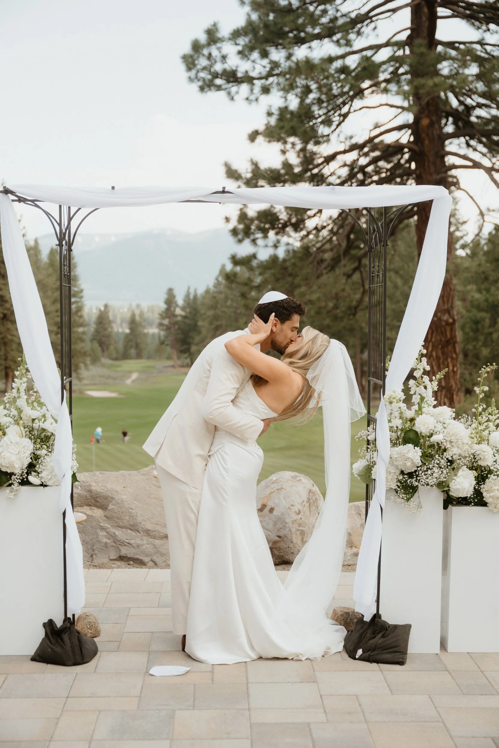 A bride and groom kiss under a wedding arch decorated with white fabric and flowers, set outdoors in a scenic mountain landscape.