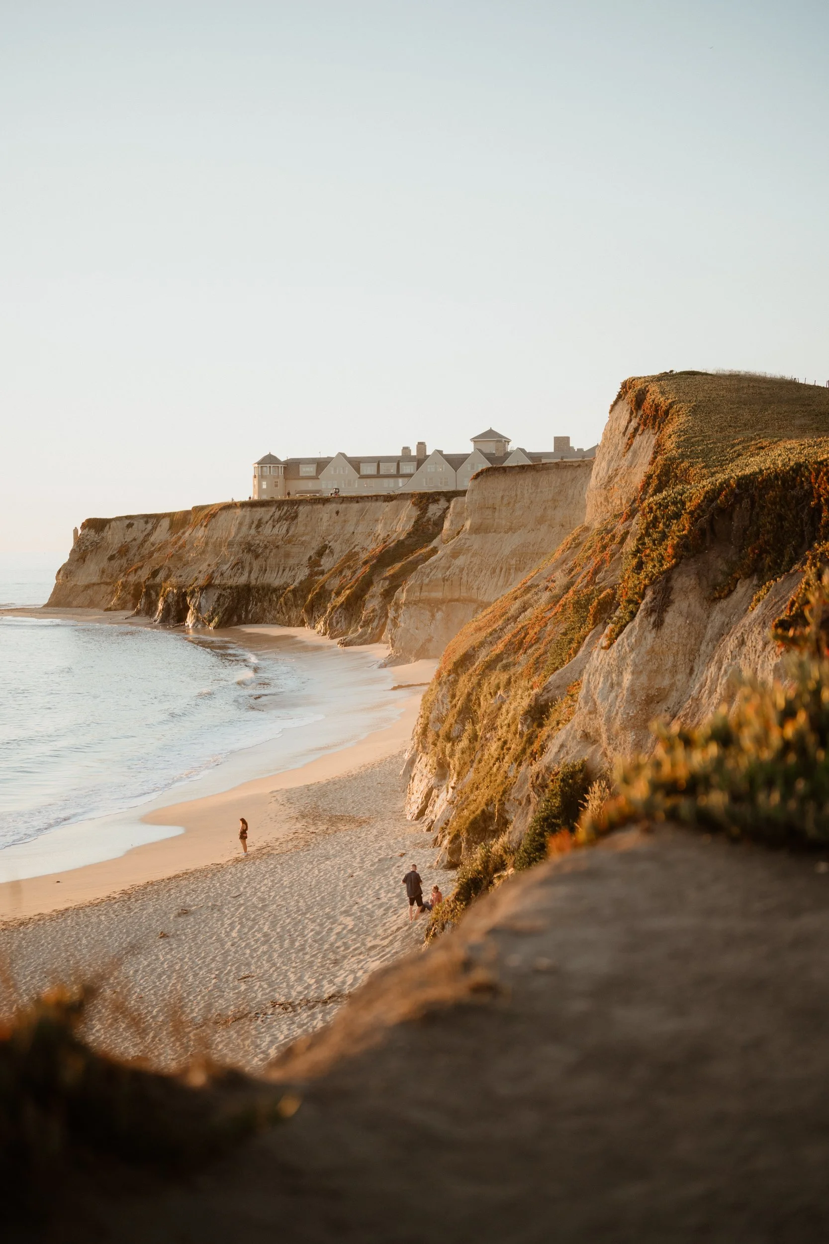 View of cliffs overlooking a sandy beach with a few people walking along the shoreline, and large houses on top of the cliffs during sunset.