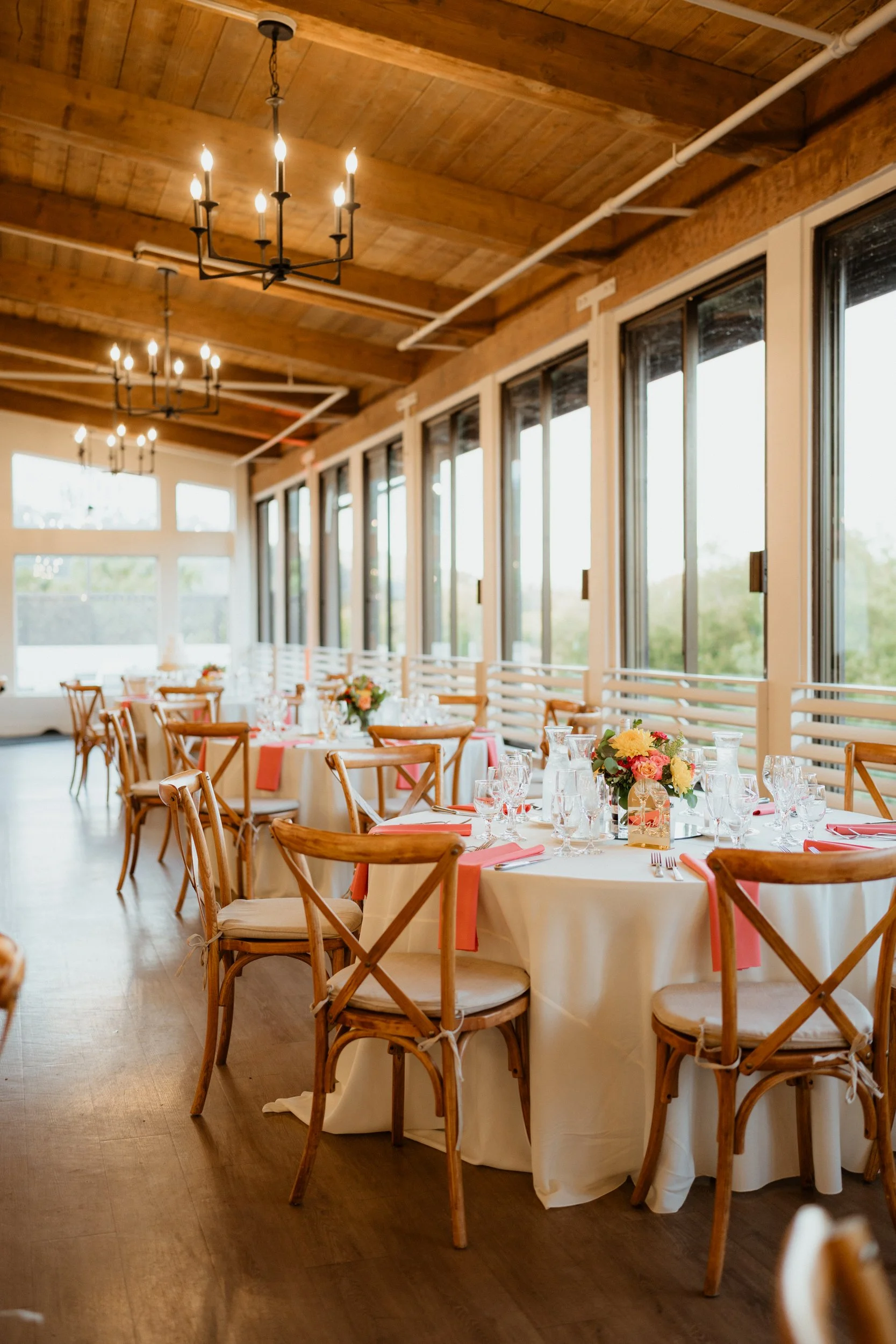 Indoor dining area with round tables covered in white tablecloths, decorated with flowers and glassware, surrounded by wooden chairs, near large windows, with wooden ceilings and chandeliers.