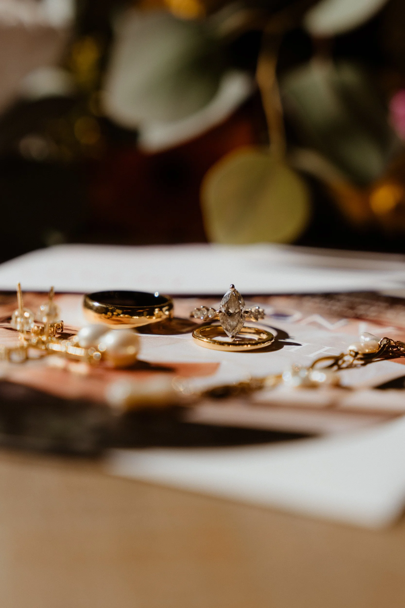 Close-up of jewelry including rings, earrings, and a bracelet displayed on a white surface with blurred greenery in the background.