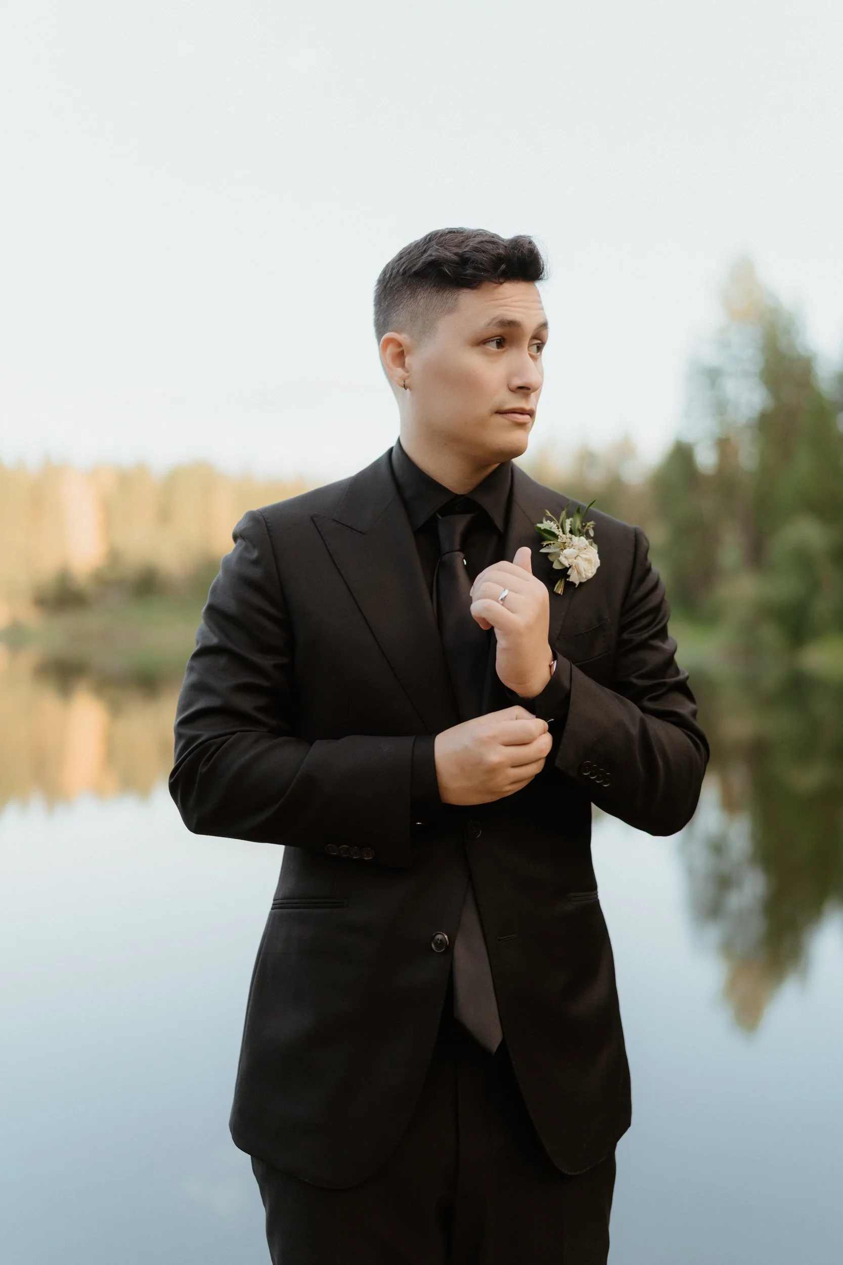 A young man dressed in a black suit standing outdoors near a body of water during daytime, with trees in the background. He has short dark hair and is adjusting his sleeve while looking to the side.