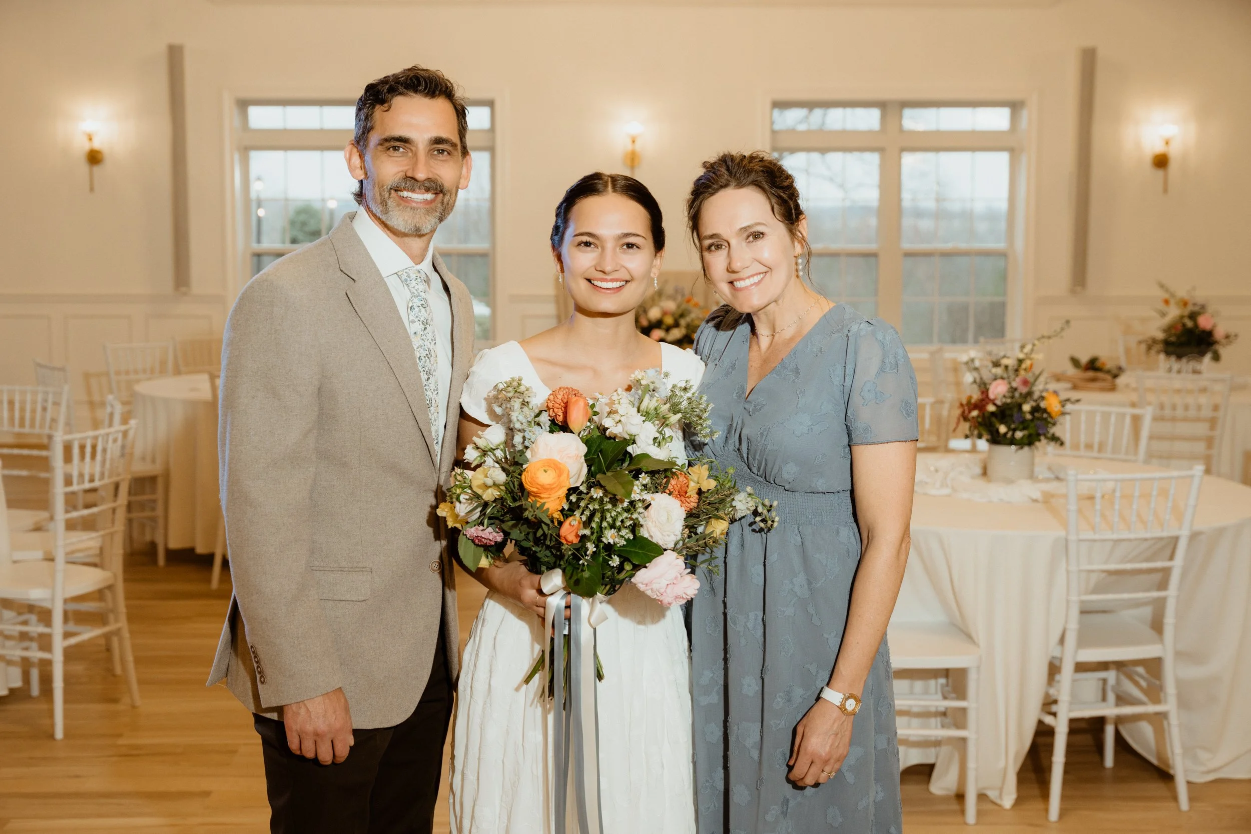 A bride in a white dress holding a colorful bouquet, standing between a man in a beige suit and a woman in a blue dress, inside a decorated reception hall with tables and flower arrangements.